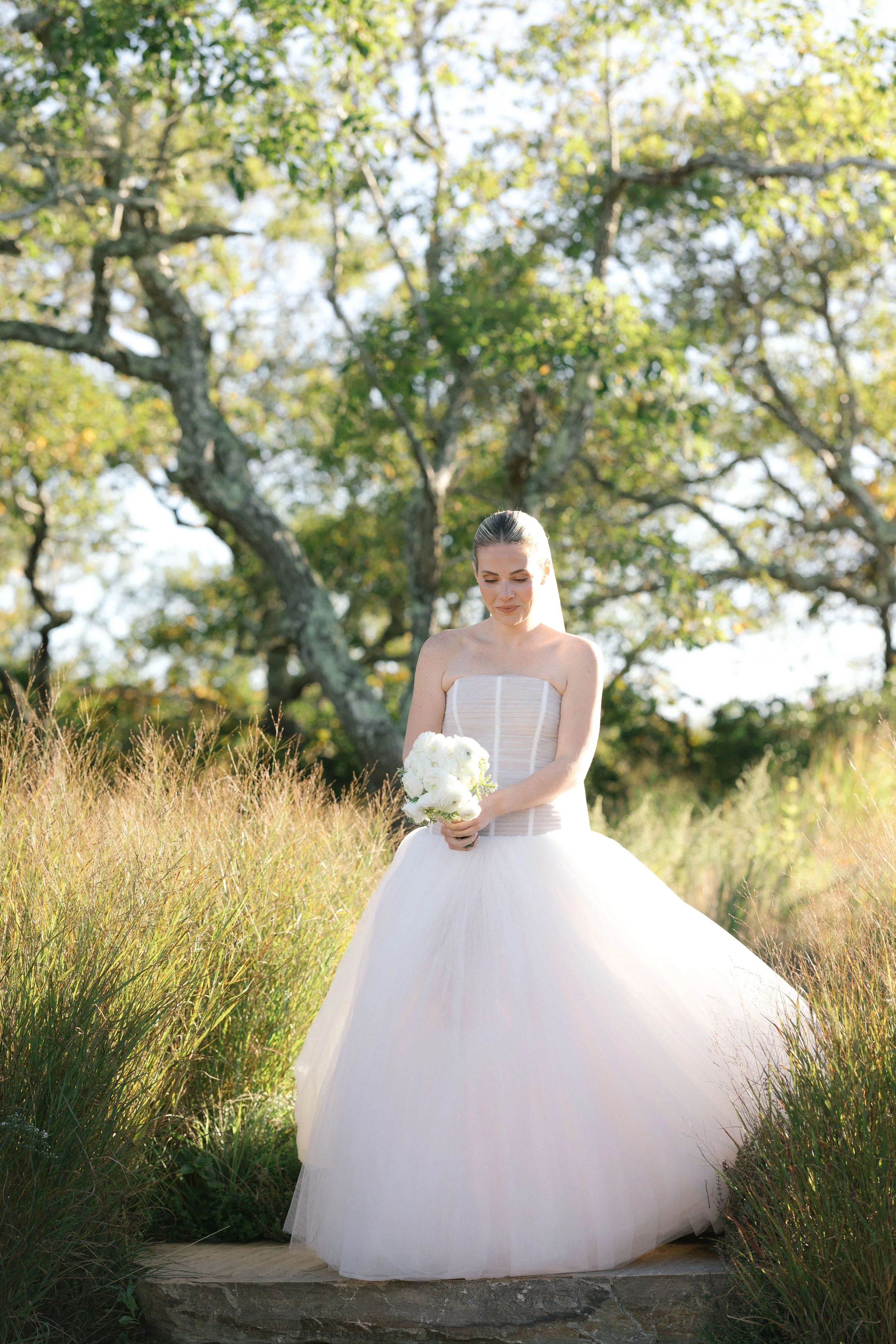 A bride in a strapless white wedding gown standing outdoors on a stone path, holding a bouquet of white flowers, with tall grass and trees in the background.