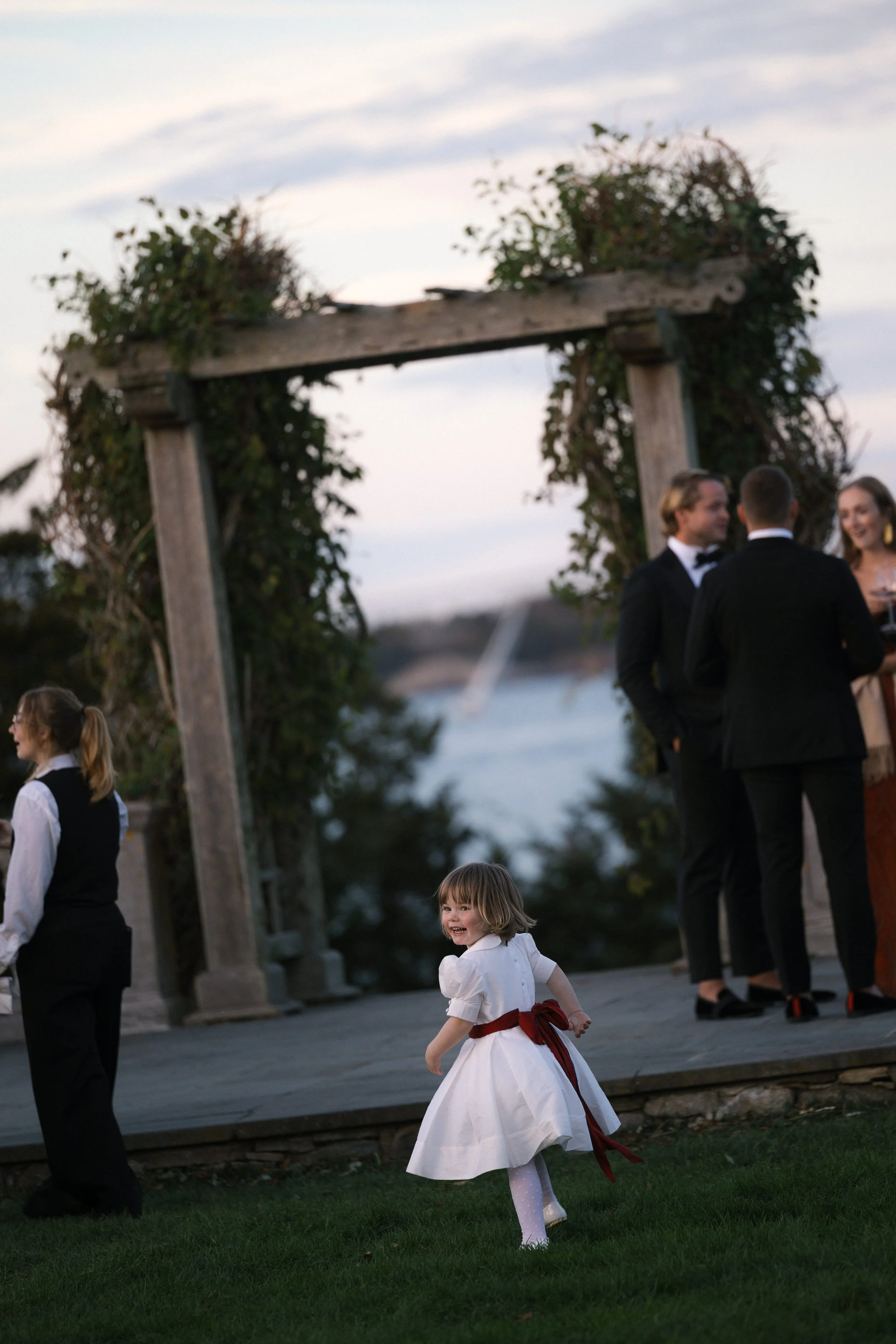 A young girl in a white dress with a red ribbon dancing on a lawn at an outdoor event during dusk, with a group of people dressed in formal attire talking near a wooden archway decorated with vines in the background.