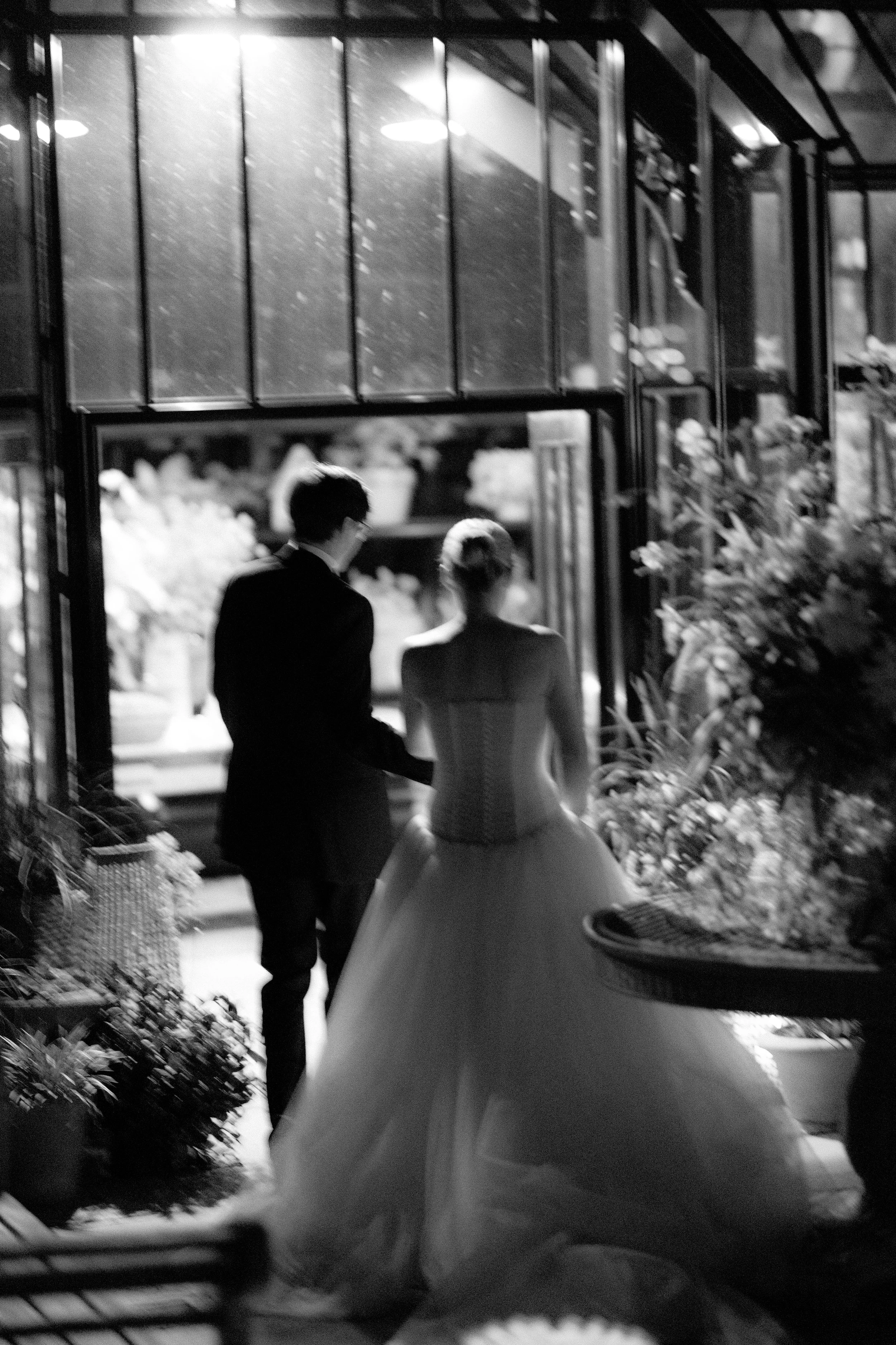 Silhouette of a couple, the man in a suit and the woman in a wedding gown, walking into a flower shop at night, with flowers on the right and glass windows behind them.