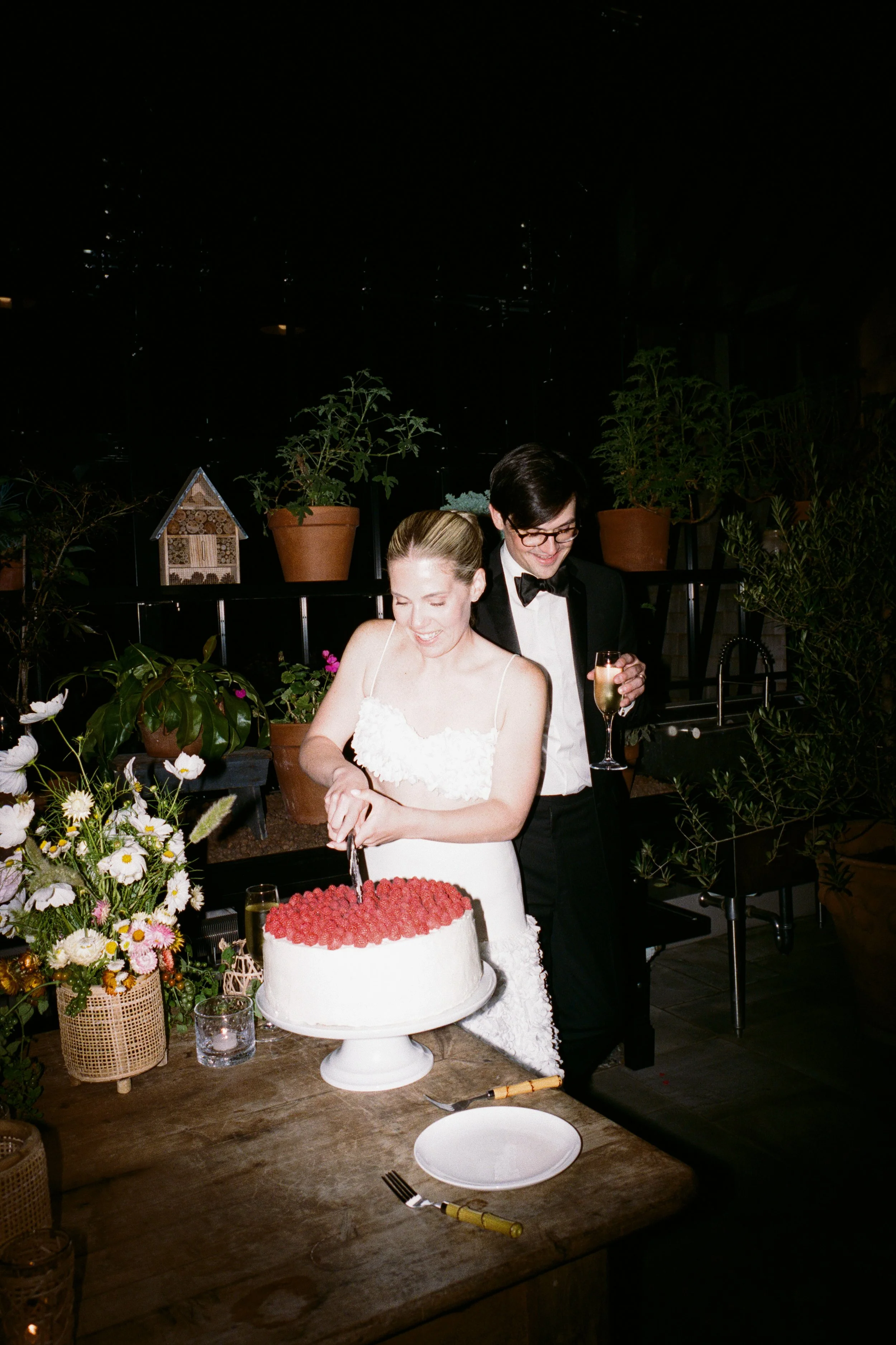 A bride and groom celebrate cutting their wedding cake. The bride is in a white dress with spaghetti straps, and the groom is in a tuxedo with a bow tie. They are smiling and holding a knife together. The cake is on a white stand and decorated with r