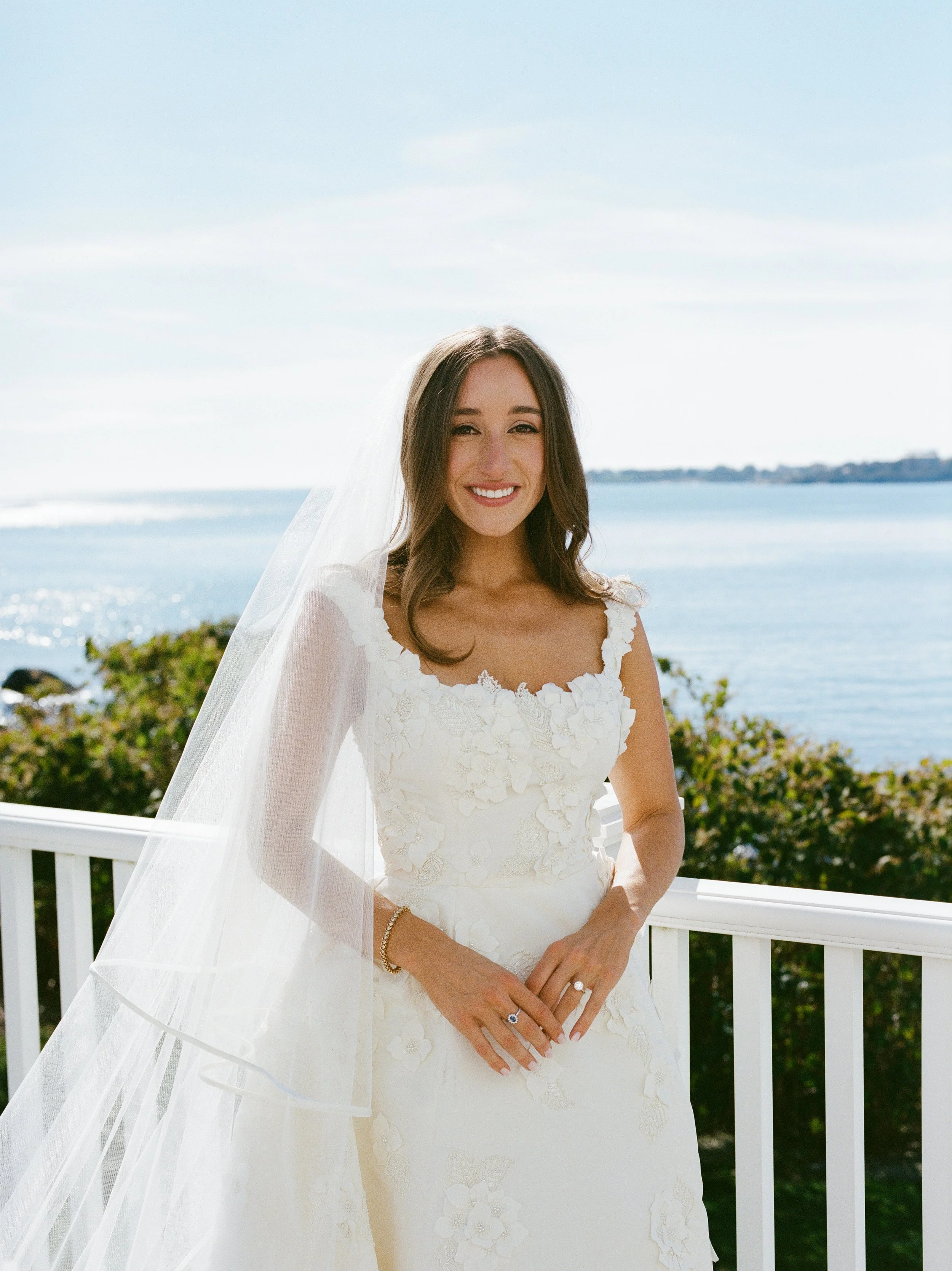 A woman in a white wedding dress with floral embroidery standing on a balcony near the ocean, smiling at the camera.
