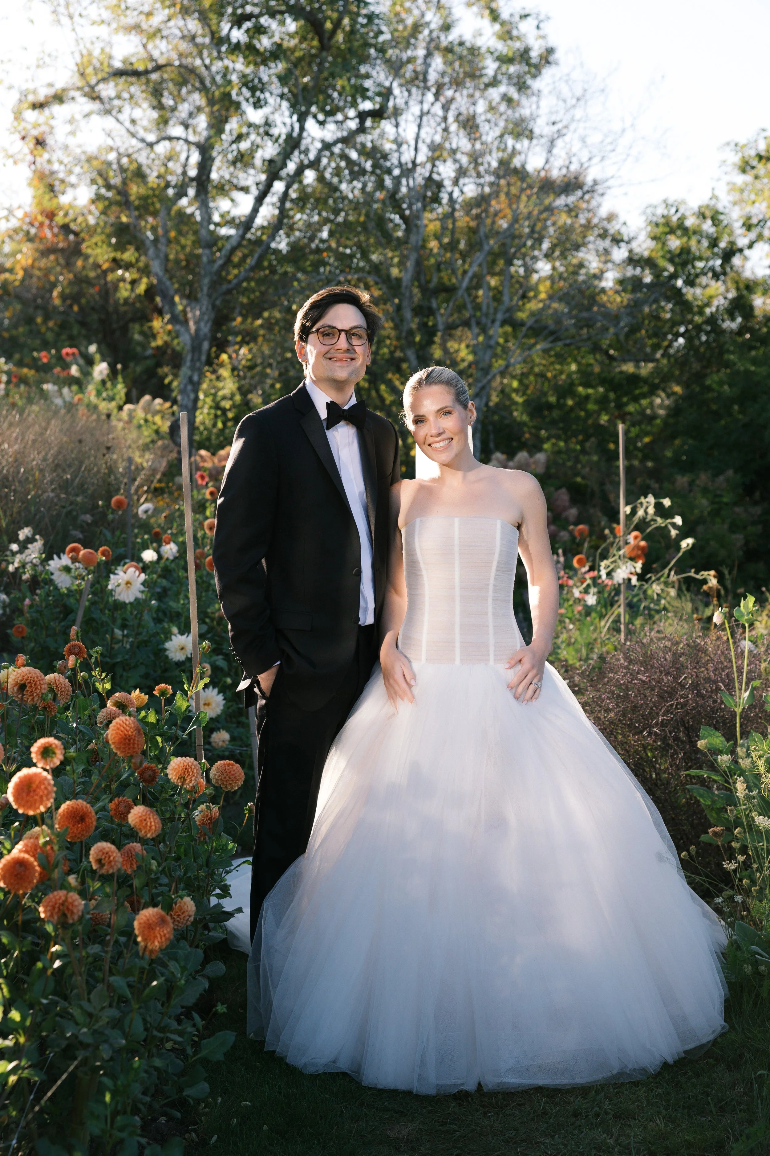 A bride and groom posing outdoors surrounded by flowers and trees, with sunlight in the background.