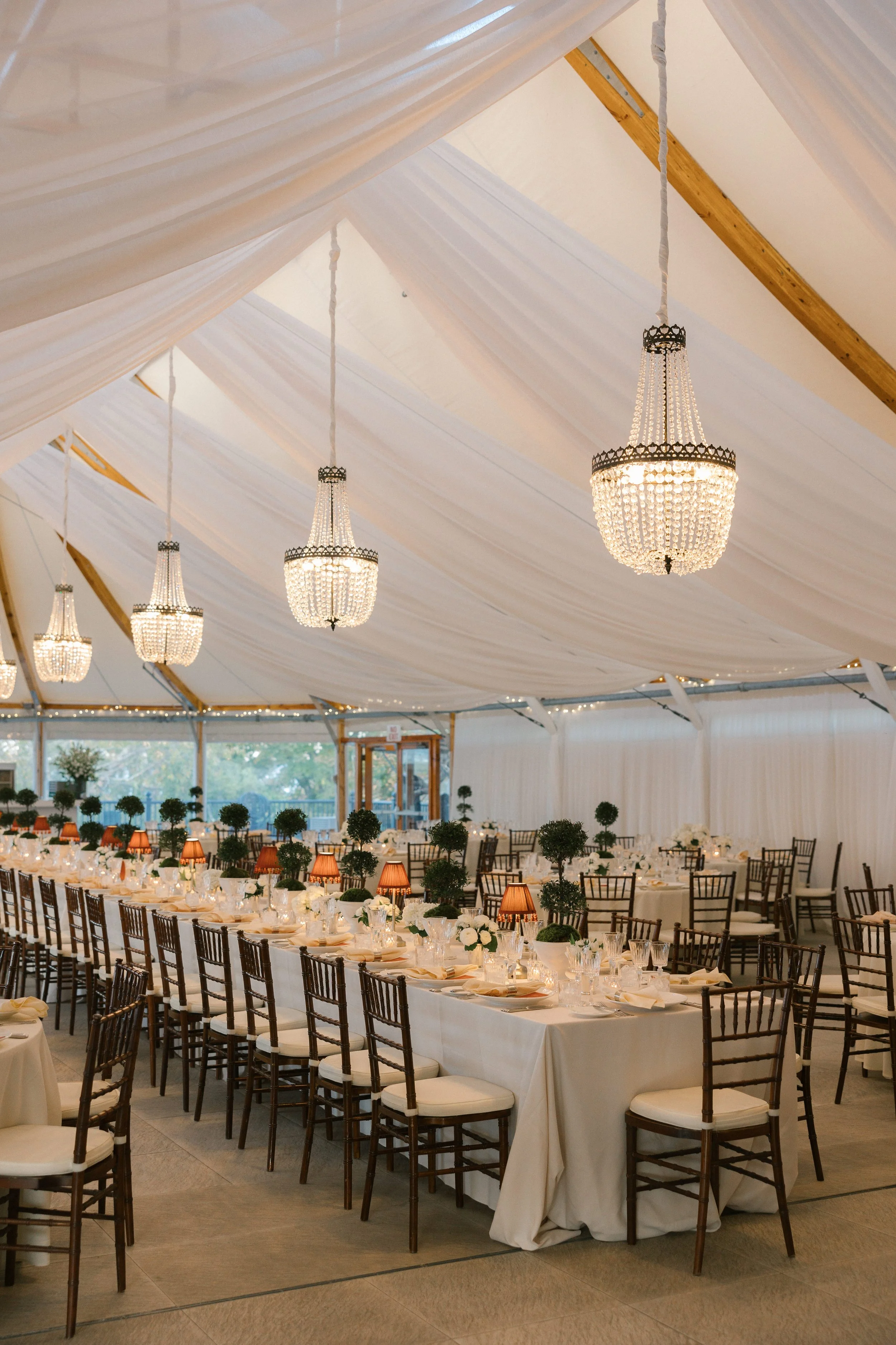 Elegant banquet hall with a long rectangular table, white tablecloth, numerous place settings, small decorative lamps, and greenery centerpieces. Crystal chandeliers hung from a tented ceiling with draped fabric.