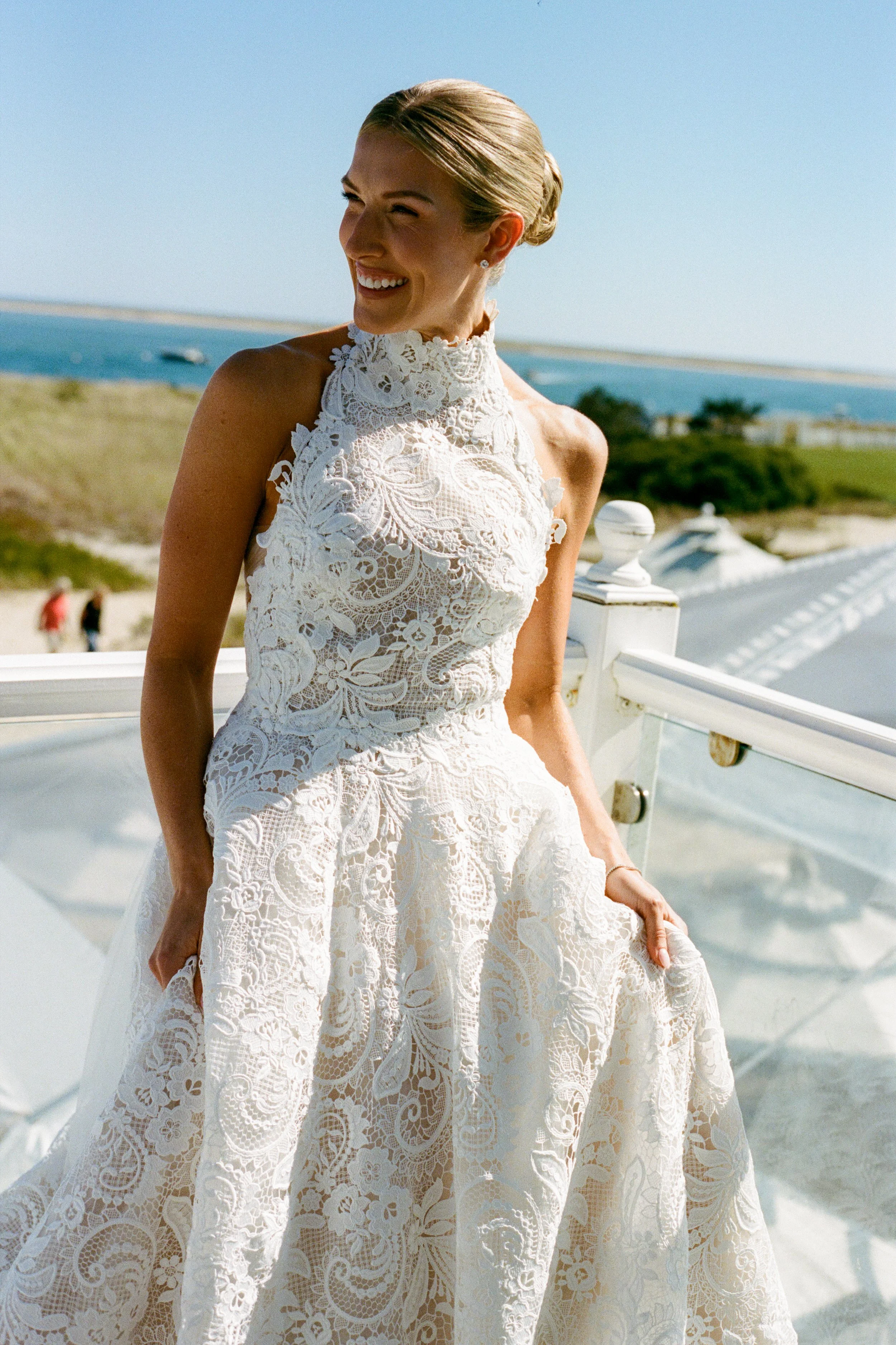 A woman in a white lace wedding dress standing outdoors on a bright, sunny day with a scenic ocean and sky background.