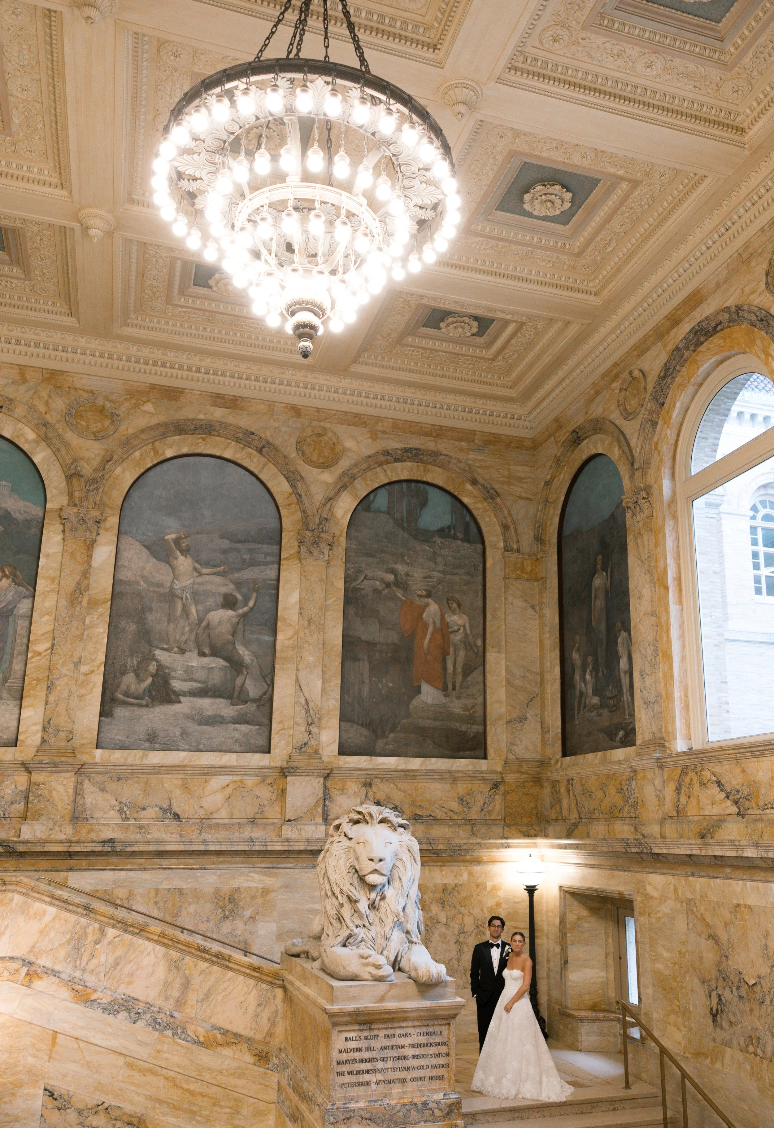 A bride and groom in formal wedding attire pose on marble stairs inside a grand museum or palace with ornate ceiling, large chandelier, marble lion sculpture, and murals on the walls.