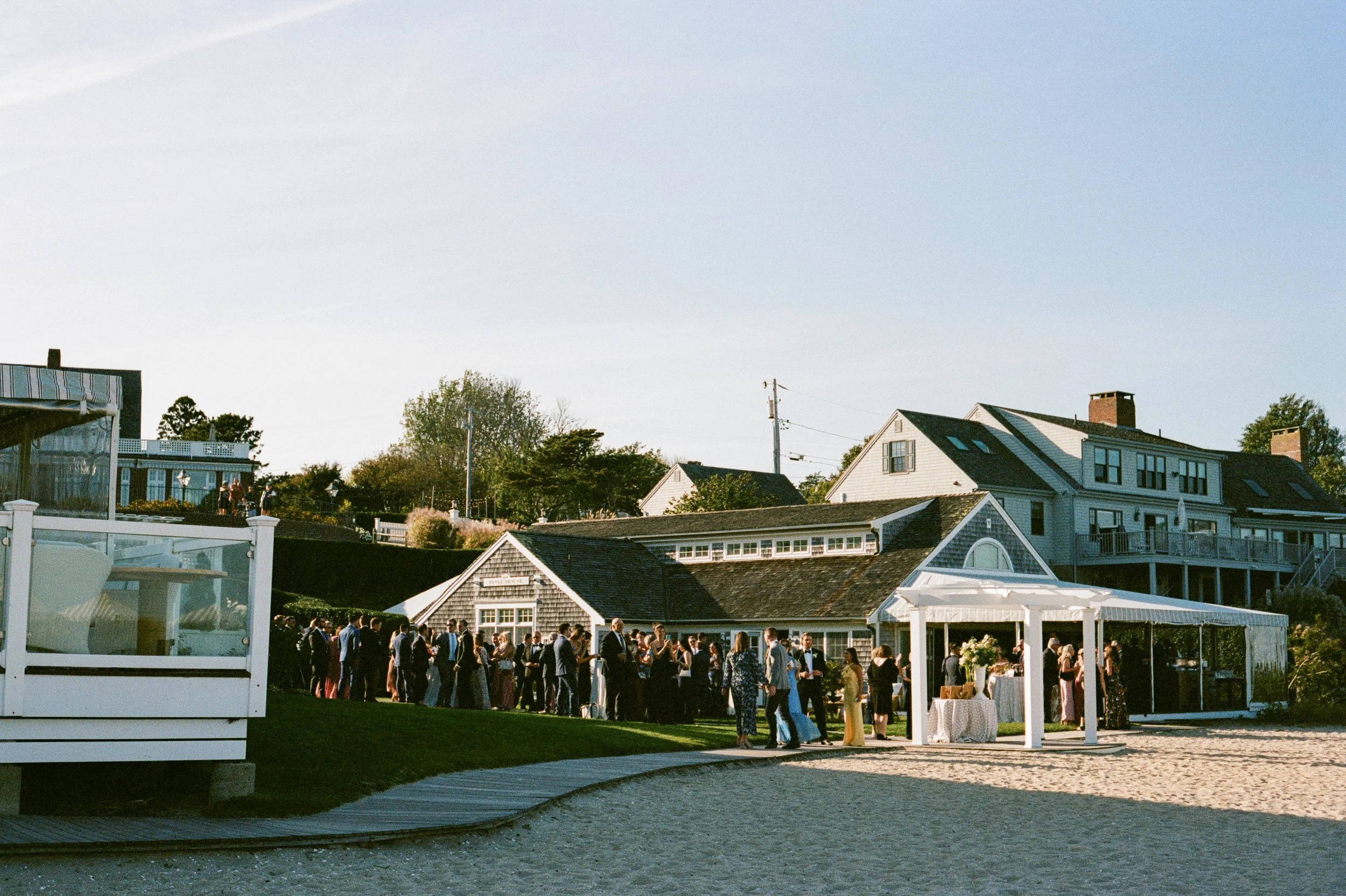 People gathered at an outdoor event under a white tent on a beachside lawn, with houses and trees in the background during late afternoon or early evening.