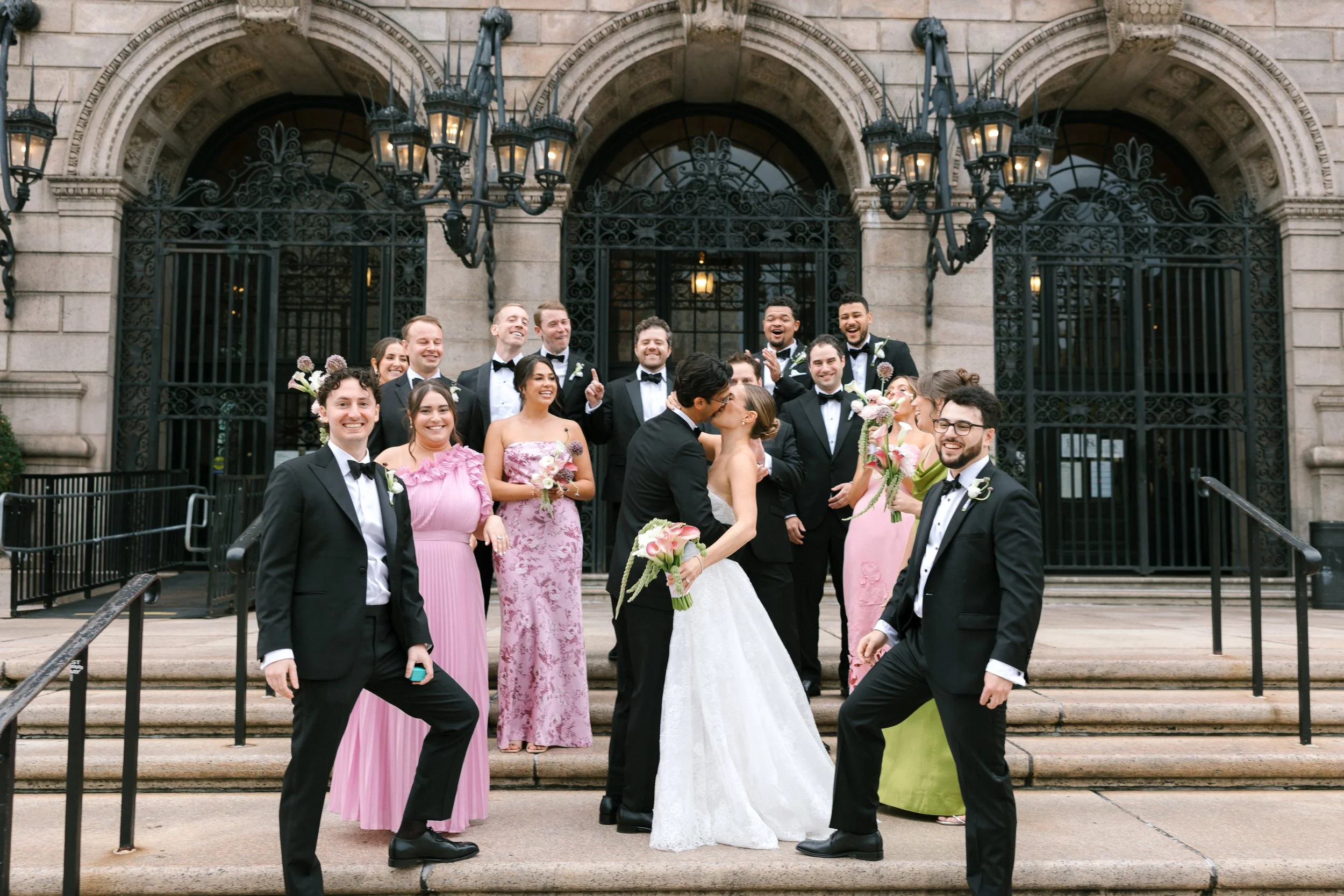 A wedding party standing on steps outside a historic building with ornate black iron gates. The group includes the bride and groom sharing a kiss, surrounded by bridesmaids in pink dresses and groomsmen in black tuxedos. Everyone is smiling and celeb
