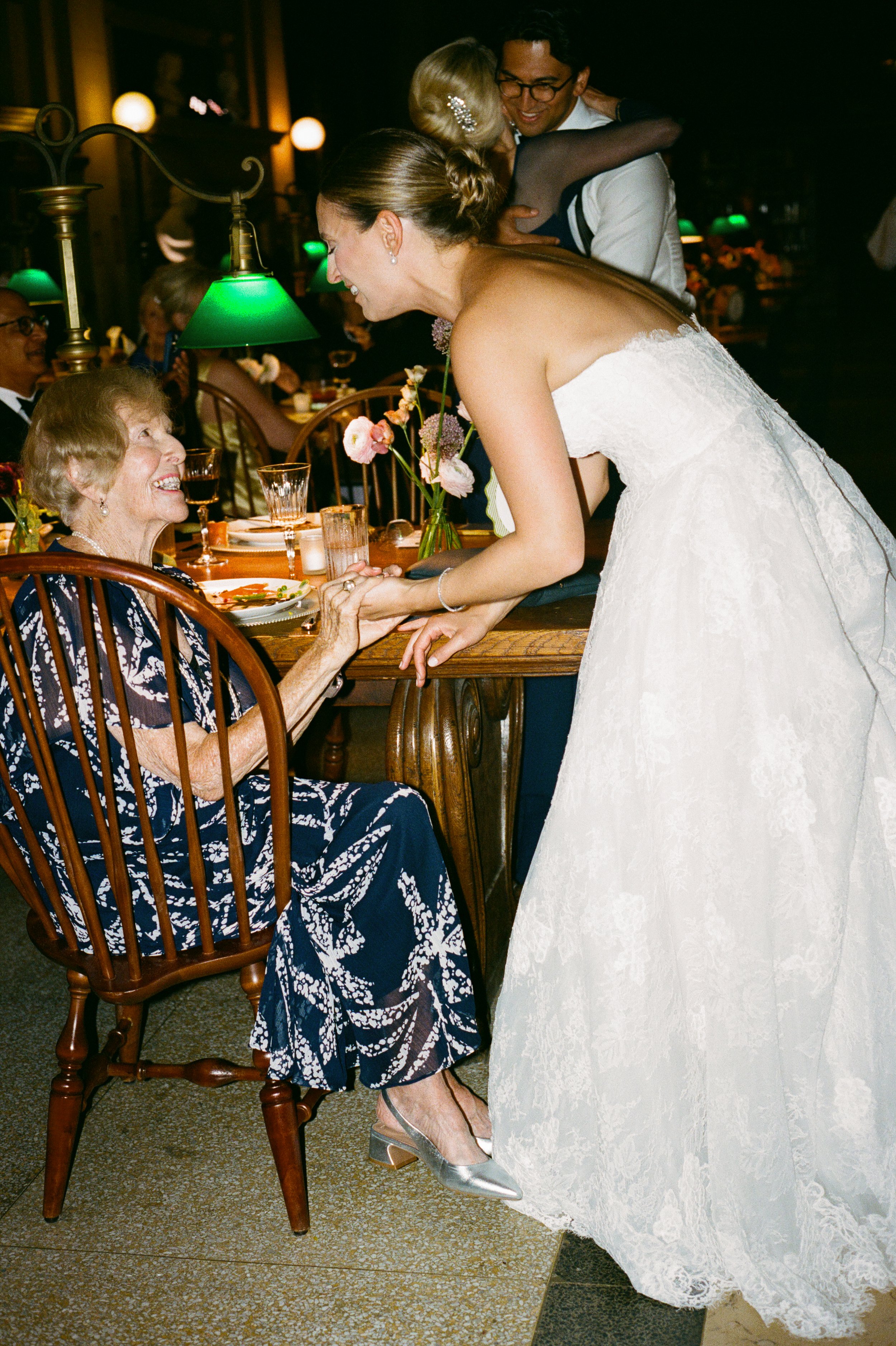 A bride in a white wedding dress joyfully interacting with an elderly woman seated at a dining table during a celebration, with other guests in the background.
