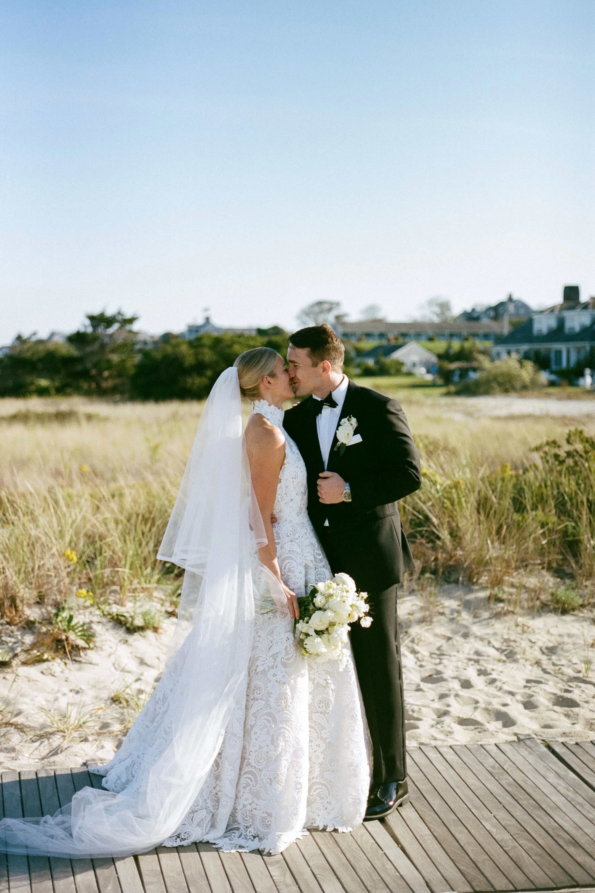Bride and groom sharing a kiss on a beach during their wedding, with the bride holding a bouquet of white flowers and wearing a lace wedding dress, the groom in a black tuxedo, and a clear blue sky in the background.