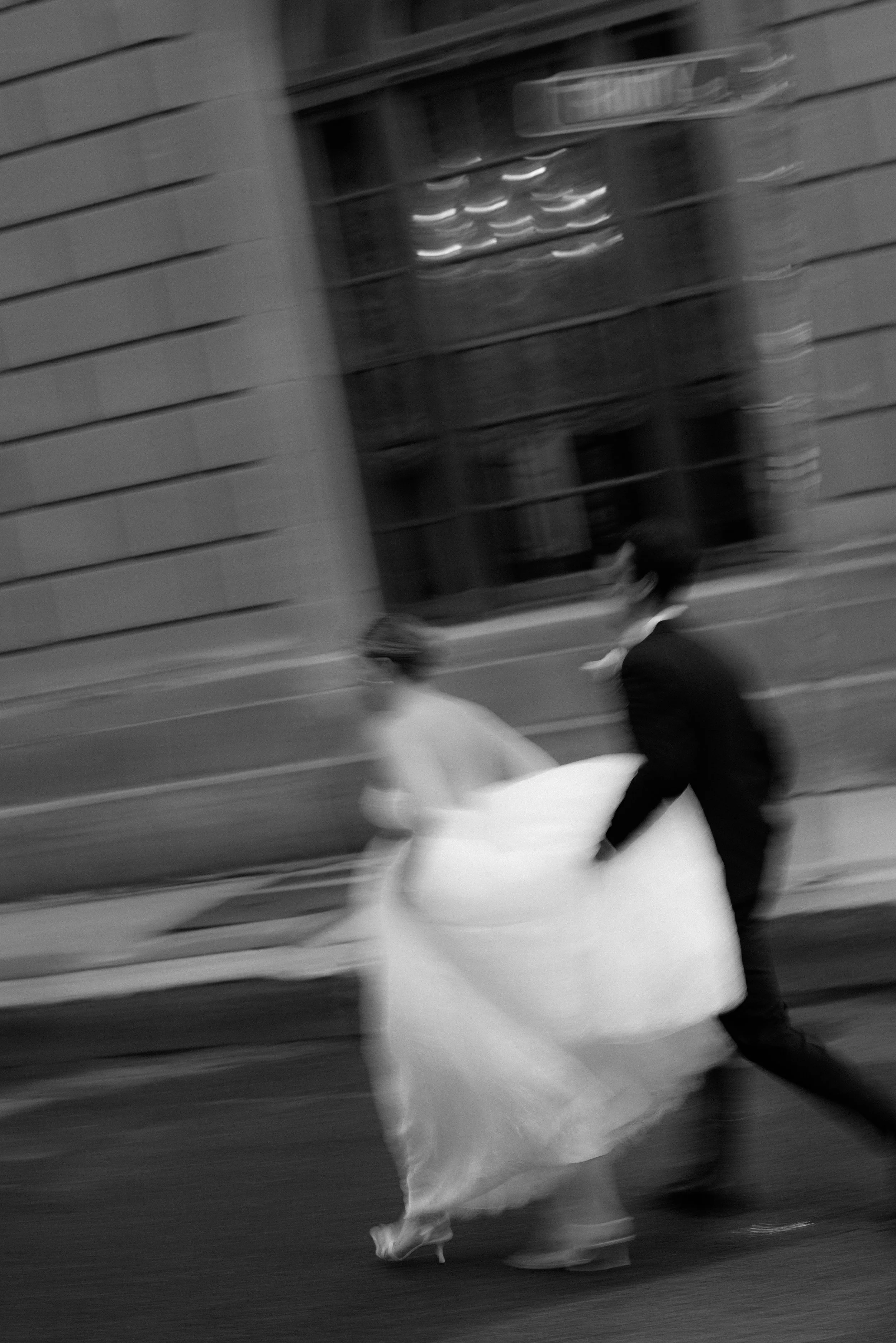 A black and white photo of a man and woman holding a wedding dress together on a city street, with motion blur.