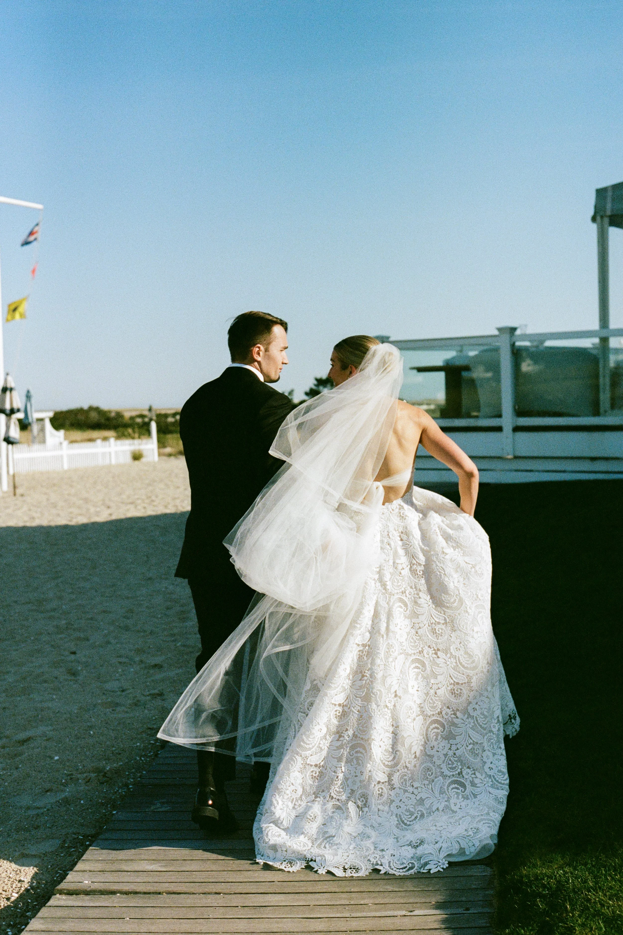 Bride and groom walking on a wooden pathway at the beach, dressed in wedding attire, with the bride's veil flowing in the wind.