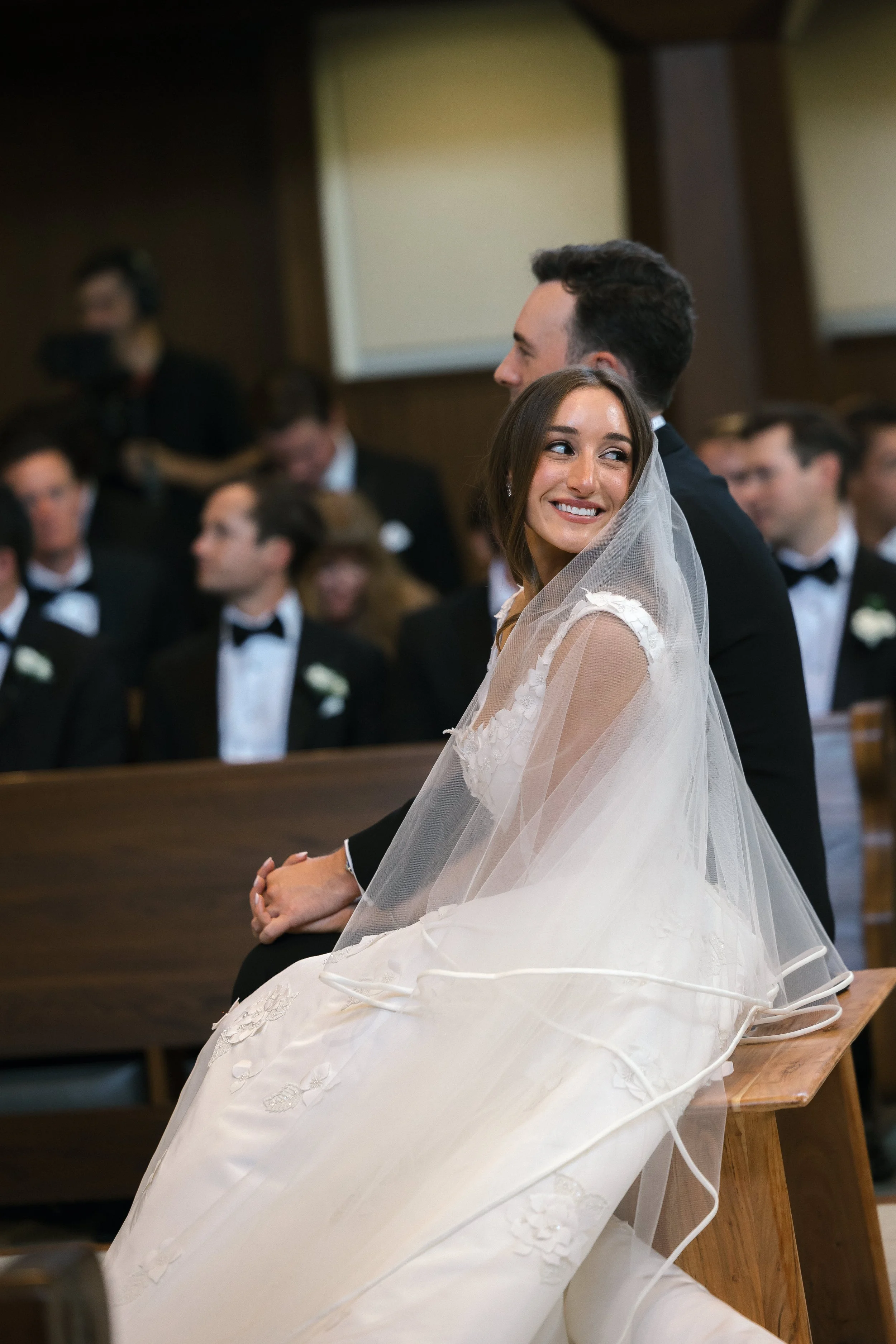 A bride smiling while holding hands with a groom during a wedding ceremony in a church, with guests in formal attire in the background.
