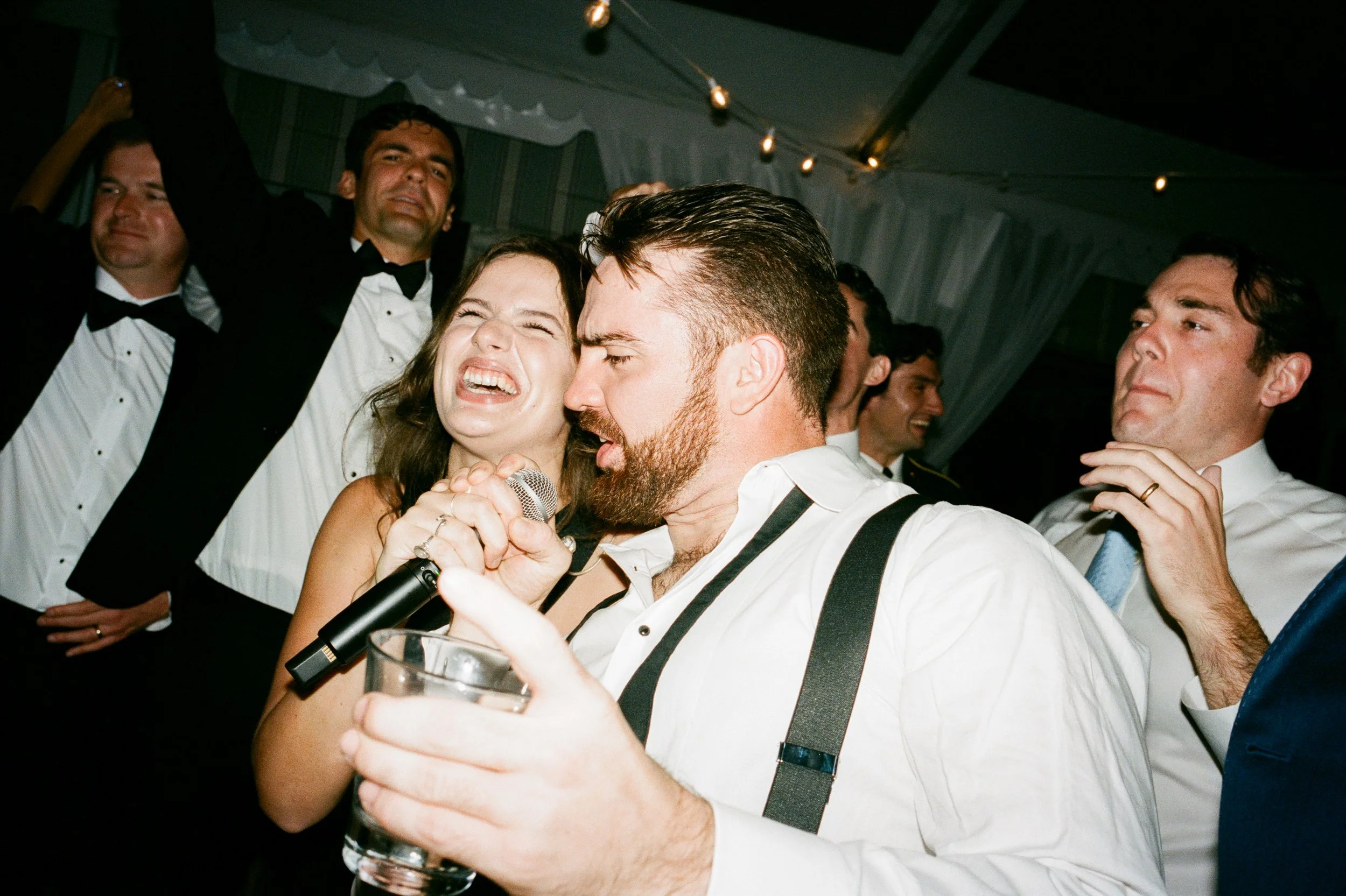 Group of people at a party singing and dancing, with some wearing formal attire and holding drinks, in a decorated indoor venue.