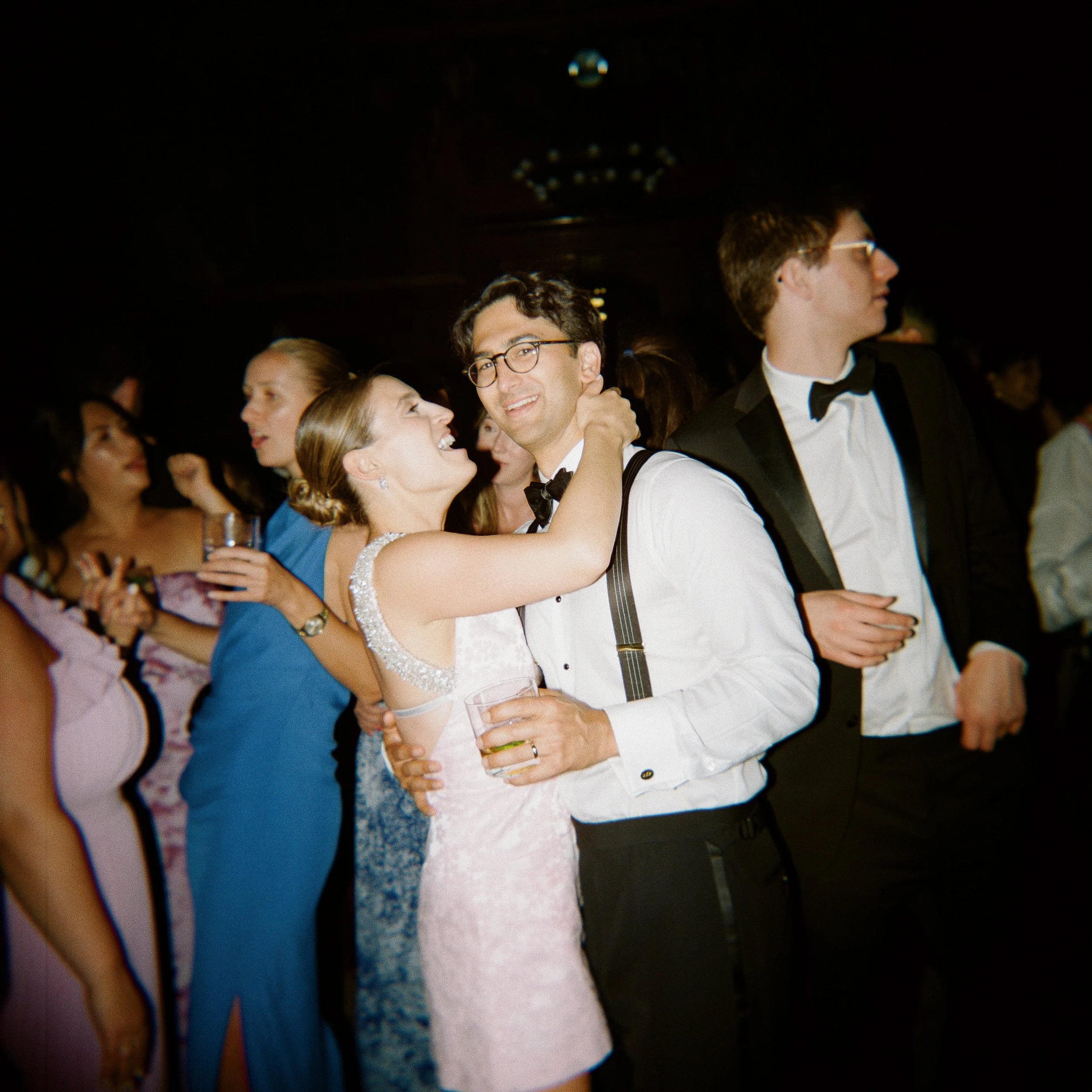 People dancing at a formal event with some wearing wedding attire, including a woman in a pink dress and a man in a white shirt with suspenders.