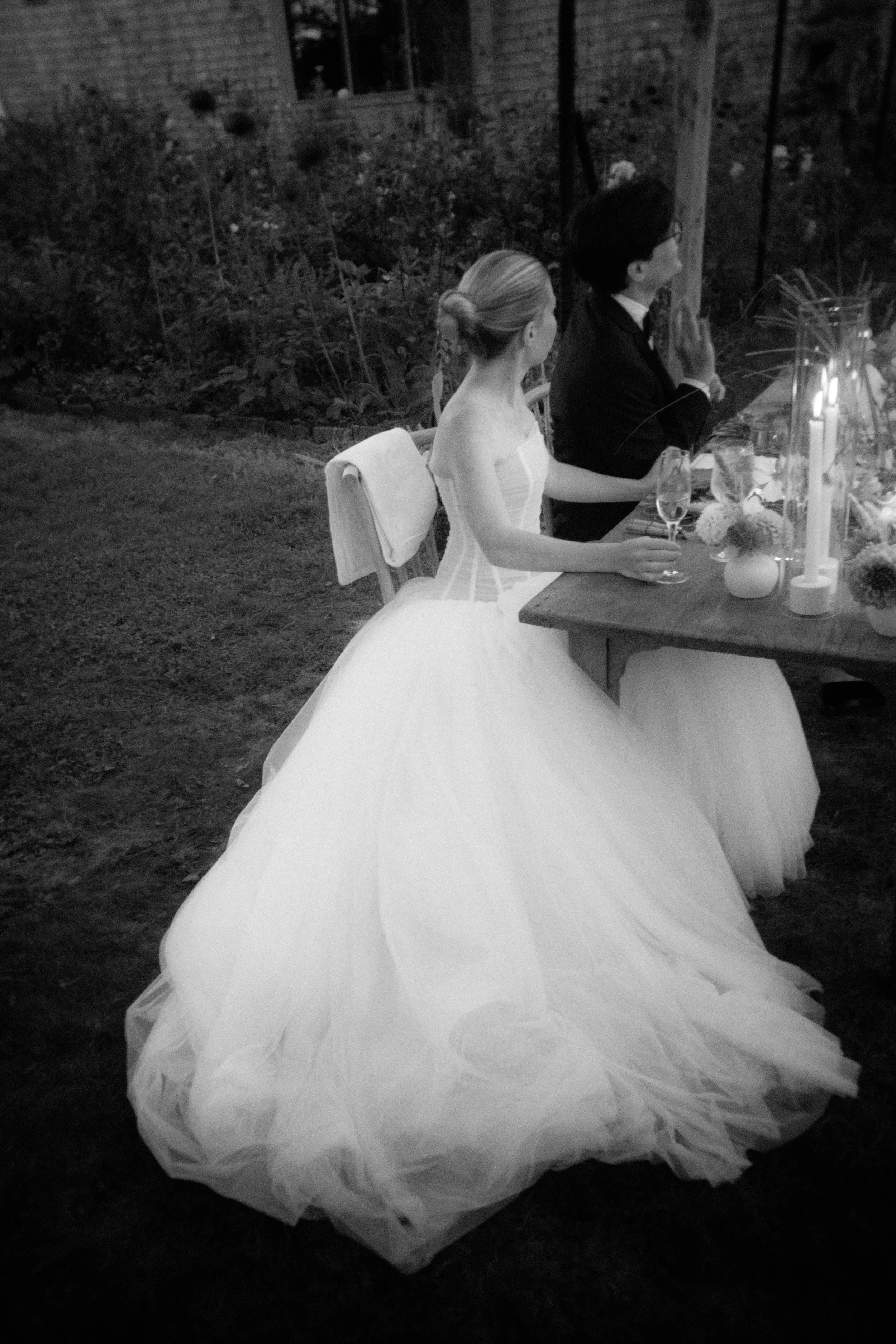 A bride in a wedding gown and a groom in a suit sit at a decorated outdoor table during a wedding reception, holding glasses and praying.