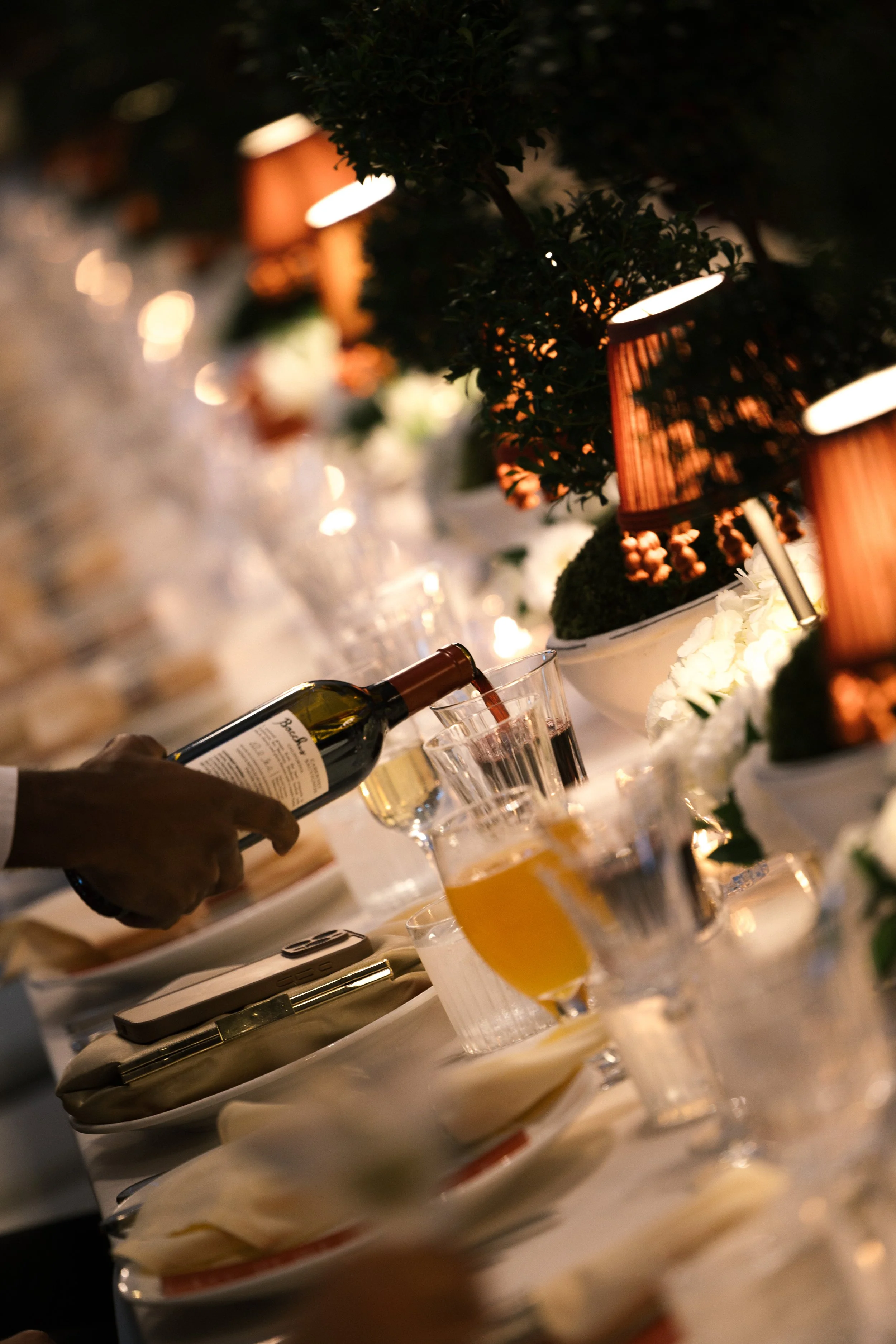 Person pouring red wine into a glass on a decorated dining table with candles and floral centerpieces.