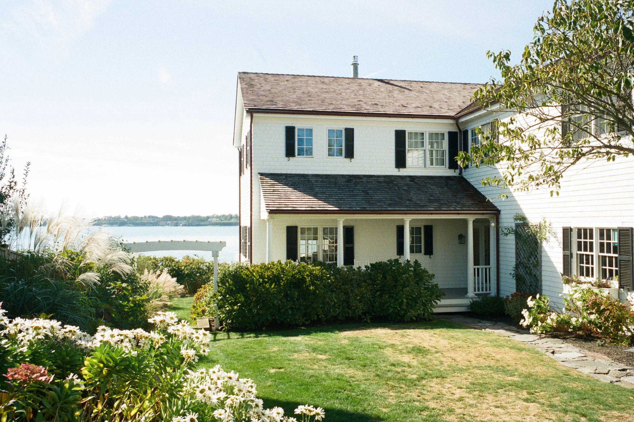 A white two-story house with black shutters, a front porch, and a garden with flowers and shrubs near a body of water.