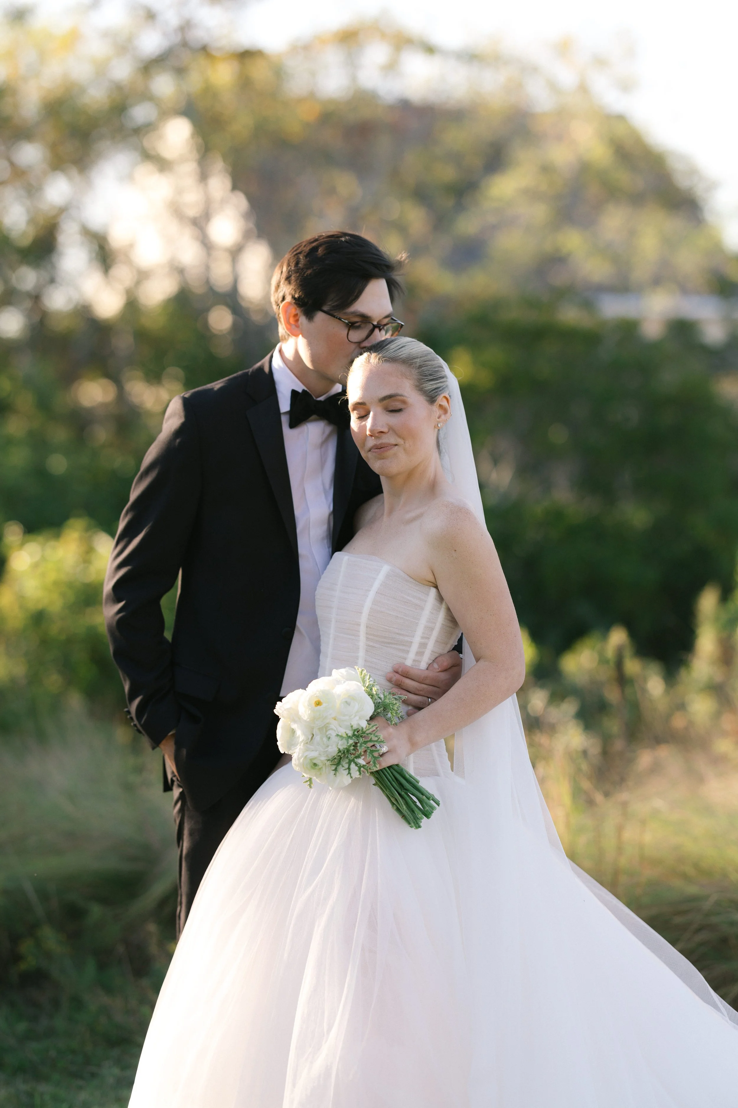 A bride in a strapless white wedding gown holding a bouquet of white flowers, standing close to a groom in a black tuxedo and glasses, outdoors with green trees and sunlight in the background.
