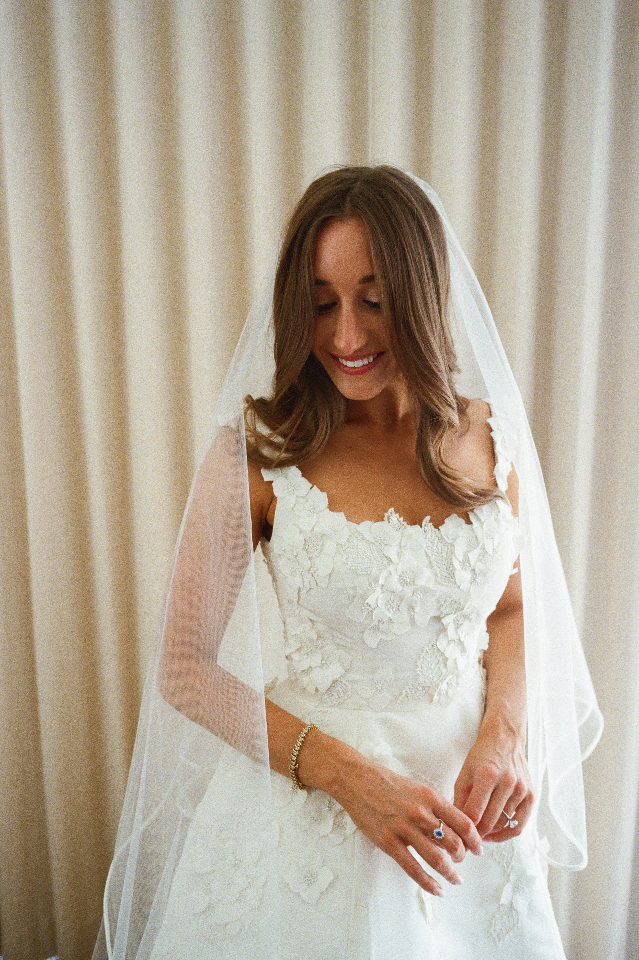 A smiling bride in a white lace wedding dress, wearing a veil, standing in front of beige curtains.