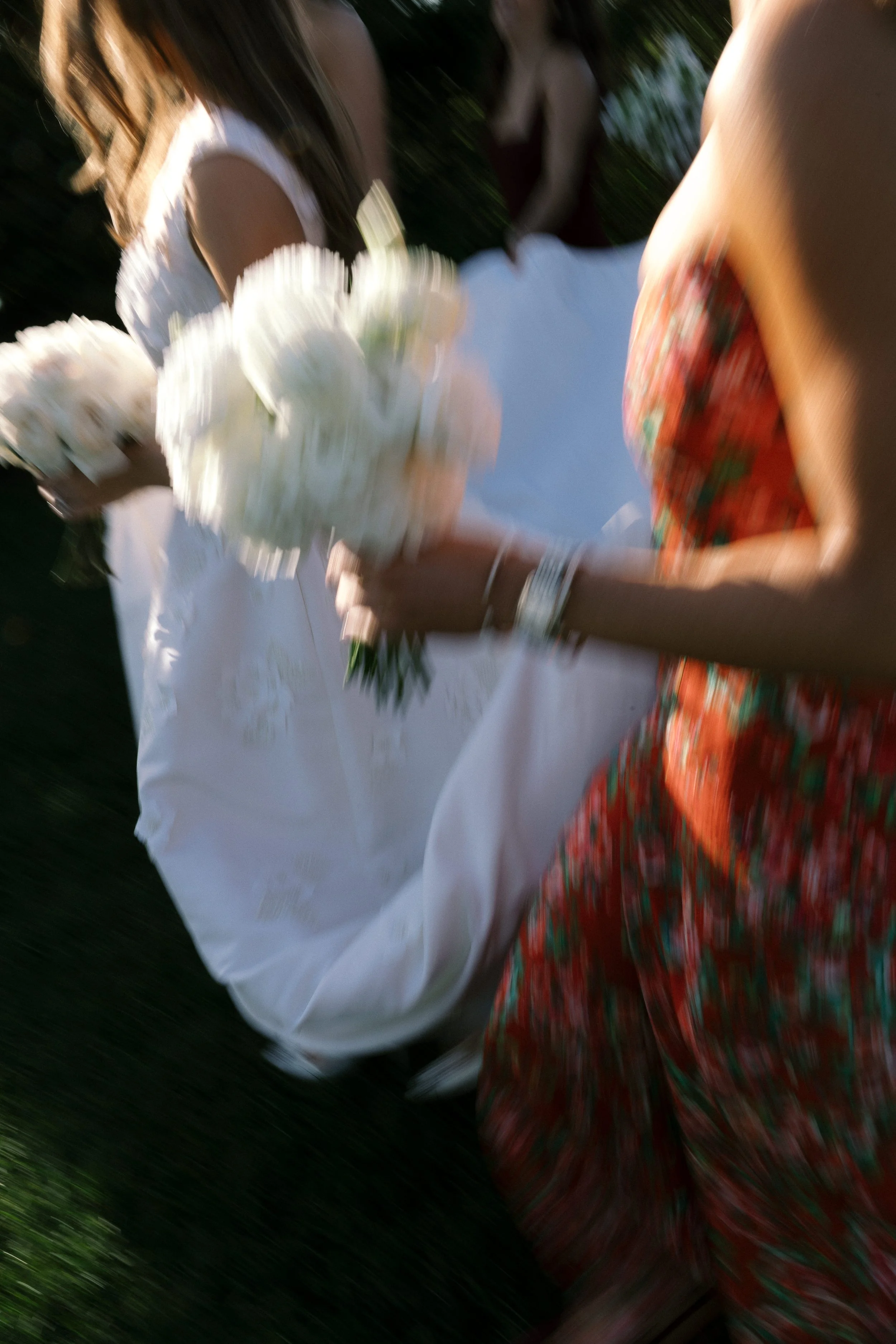 Blurred photo of a wedding scene with people, one person in a white dress holding a bouquet of white flowers, and another in a red patterned dress.