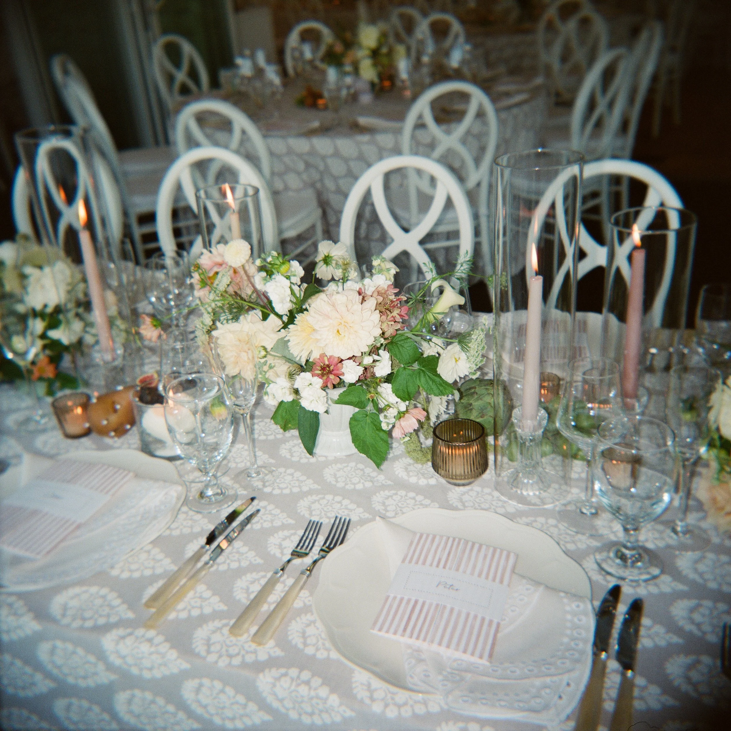 Elegant table setting at a wedding or event with a floral centerpiece, candles, glassware, and neatly arranged cutlery on a white lace tablecloth.