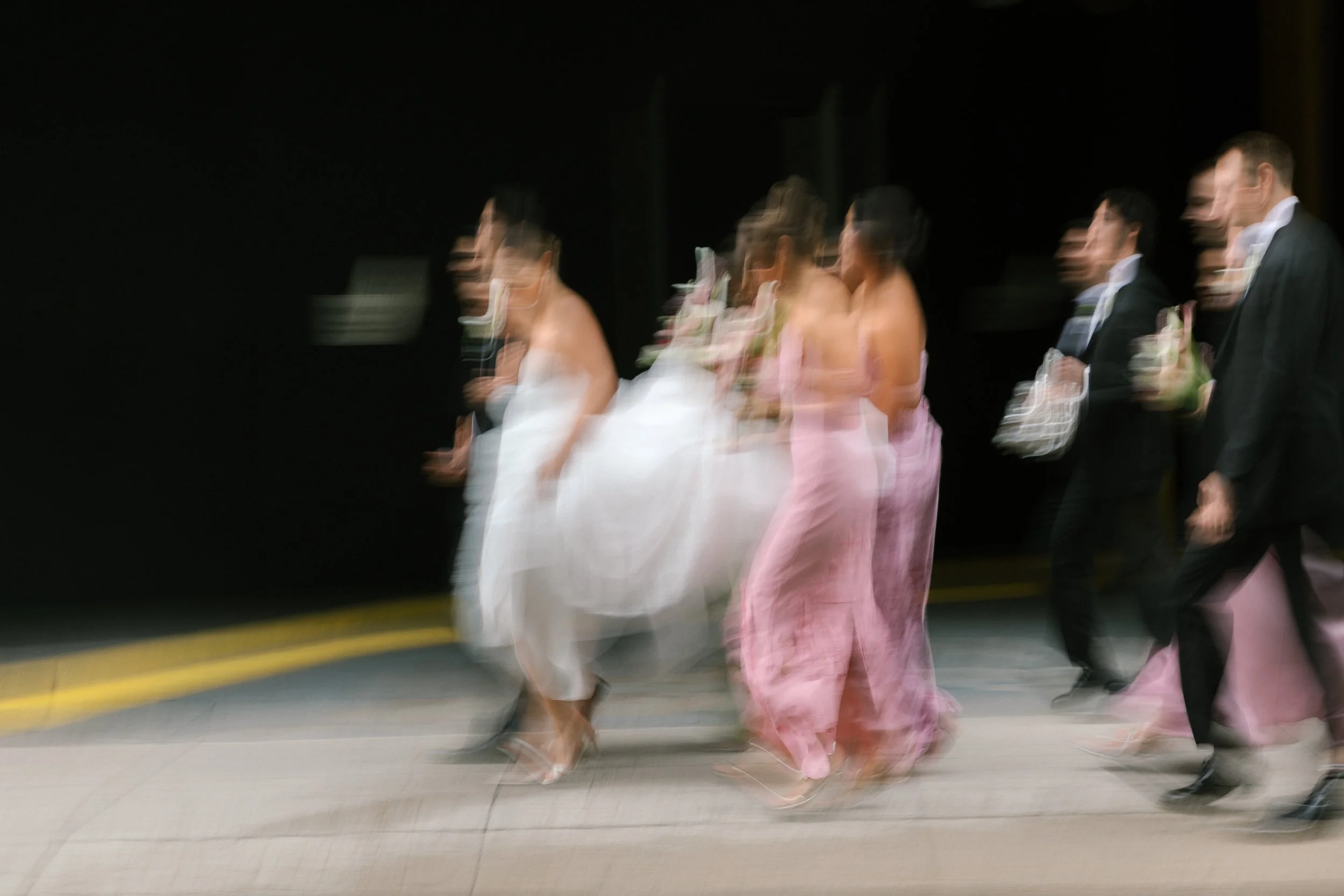 Blurred motion photo of a group of people dressed in formal attire, including women in pink and white dresses and men in black suits, walking outdoors during an evening event.