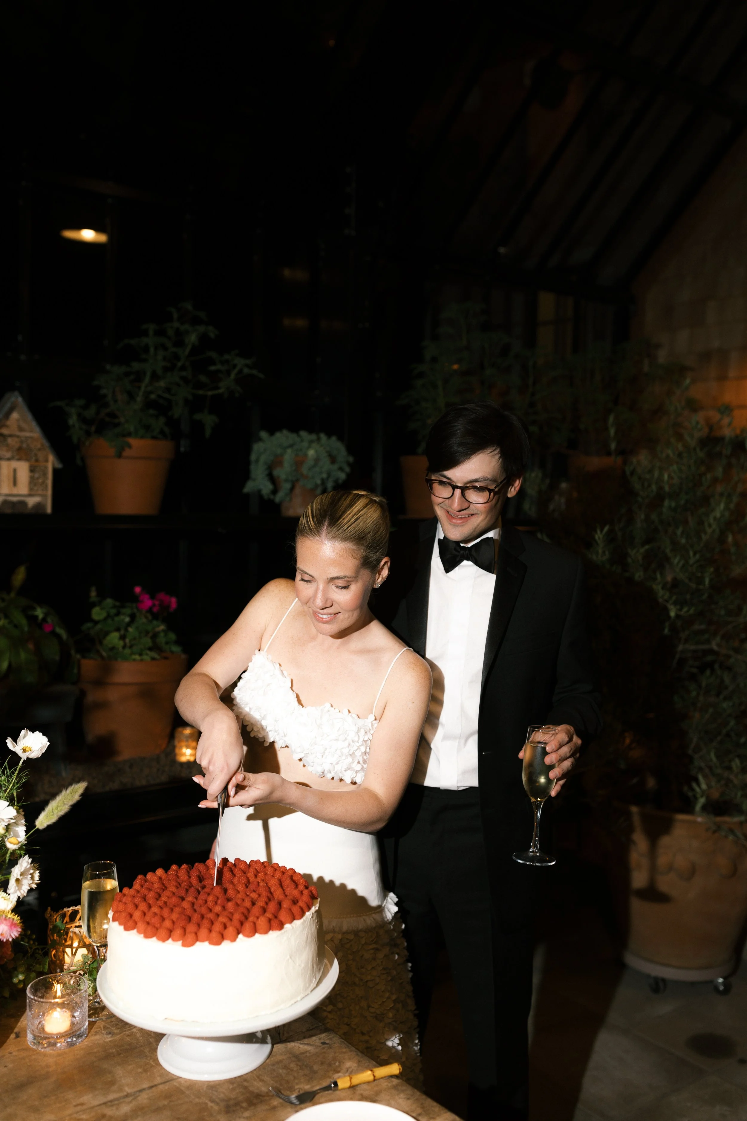 A bride in a white dress and a groom in a tuxedo celebrate at a wedding reception, cutting a cake decorated with raspberries, with glasses of champagne in hand, indoors with potted plants and candles.