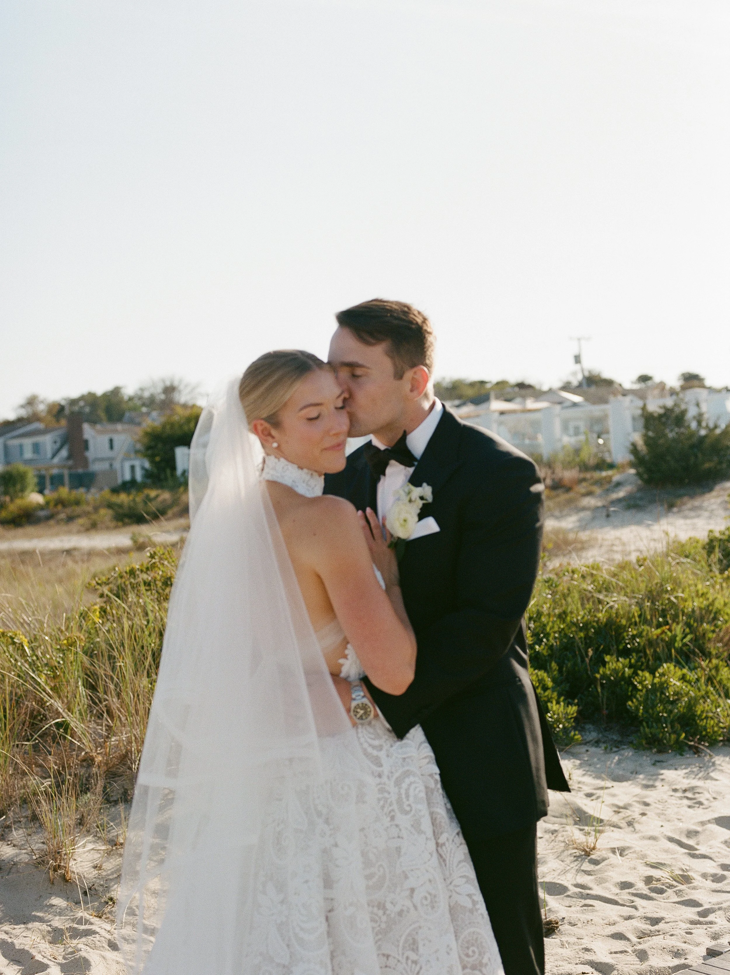 A bride and groom embrace on a beach, with the groom gently kissing the bride's forehead, during their wedding photoshoot.