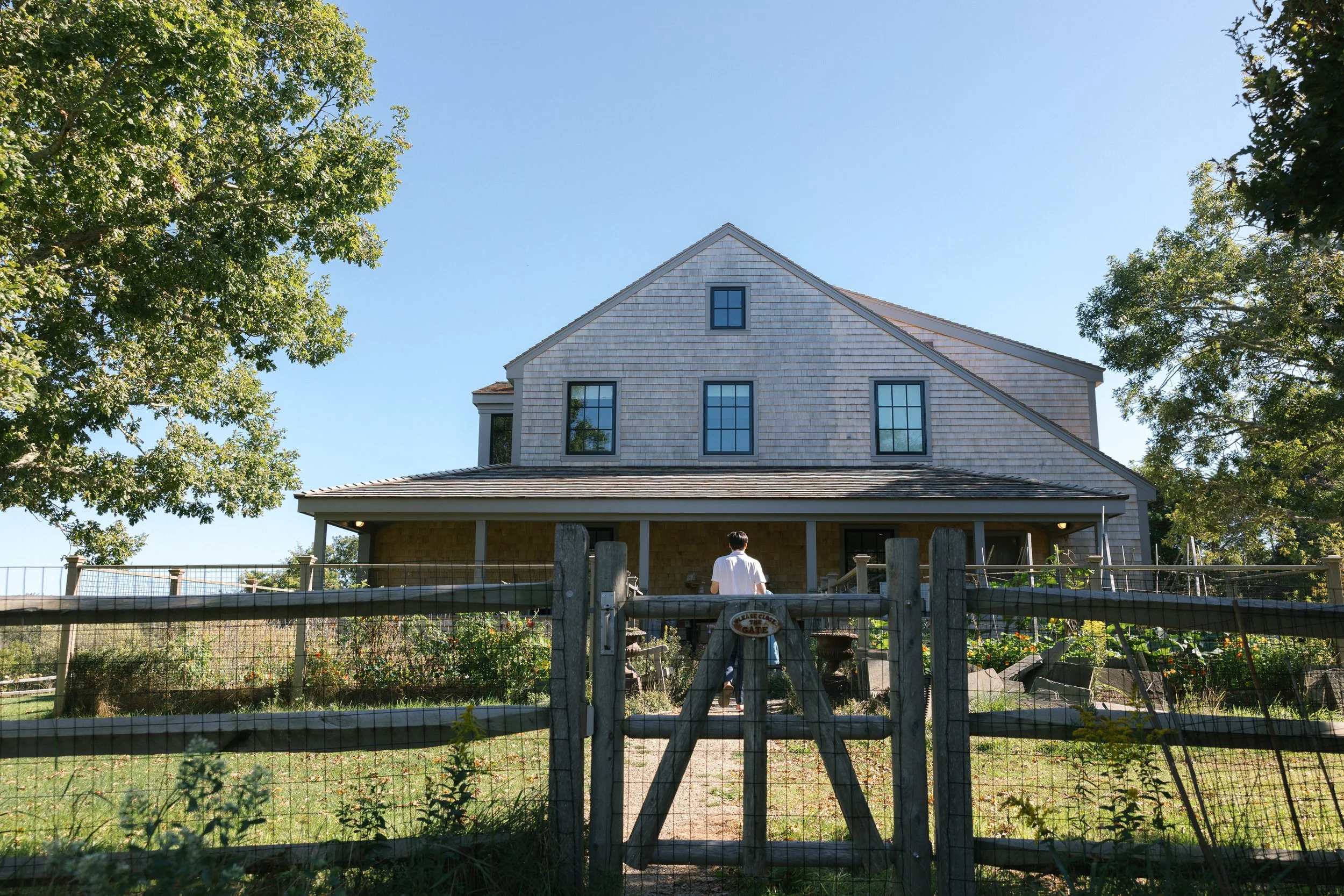 A two-story house under construction viewed through a wooden fence, with a person standing in front, on a sunny day with clear blue sky.