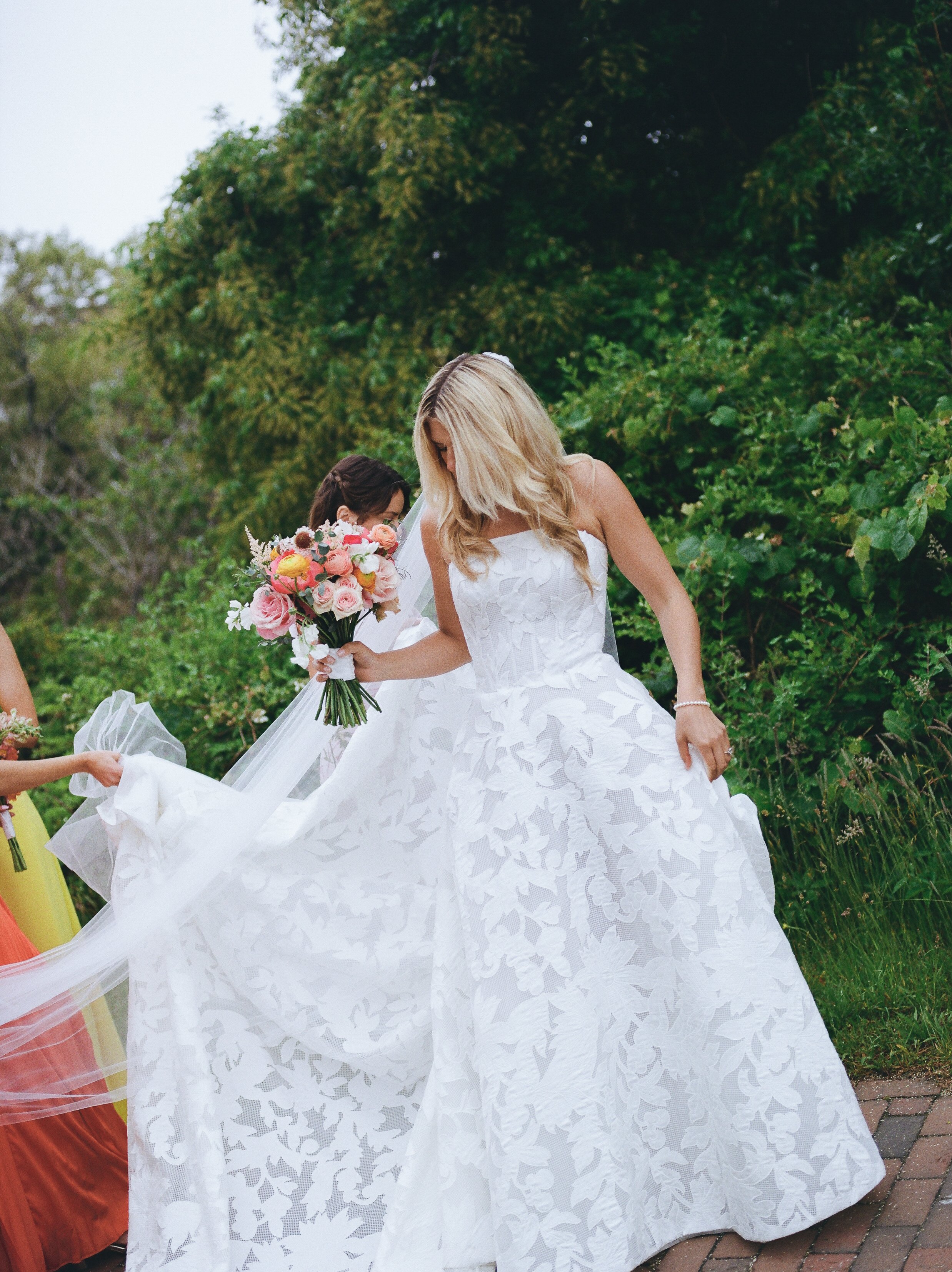 A bride in a white wedding dress holding a bouquet of pink and white flowers, standing outdoors with greenery in the background.