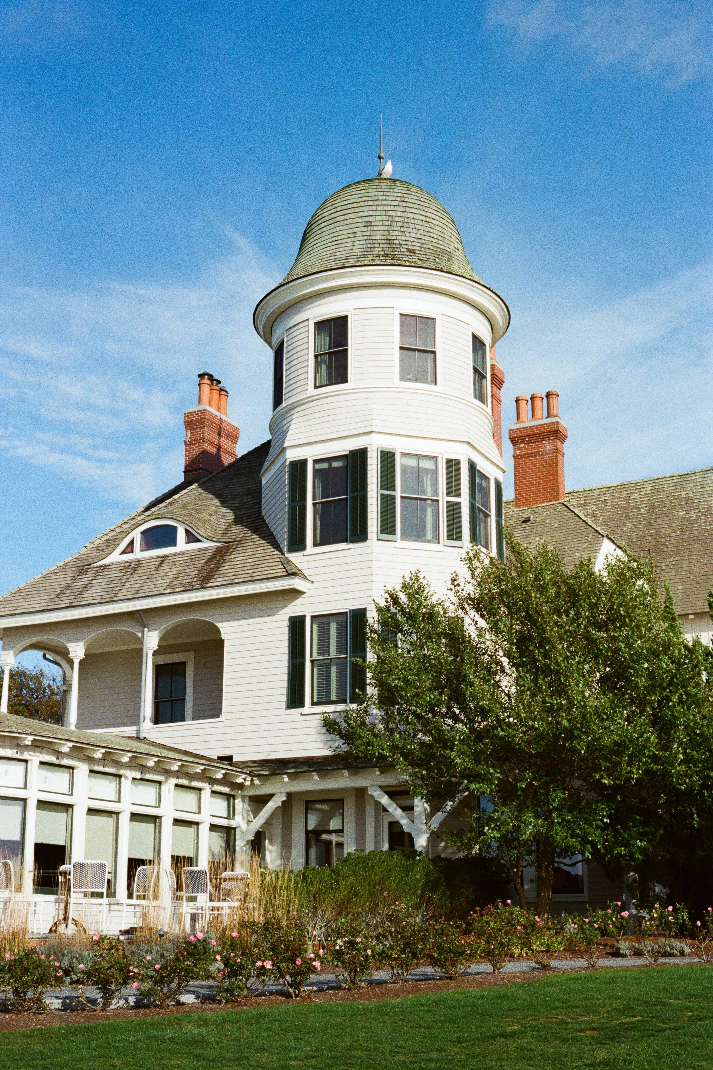 A large, historic, Victorian-style house with a prominent round tower, surrounded by a garden with bushes and flowers, under a clear blue sky.