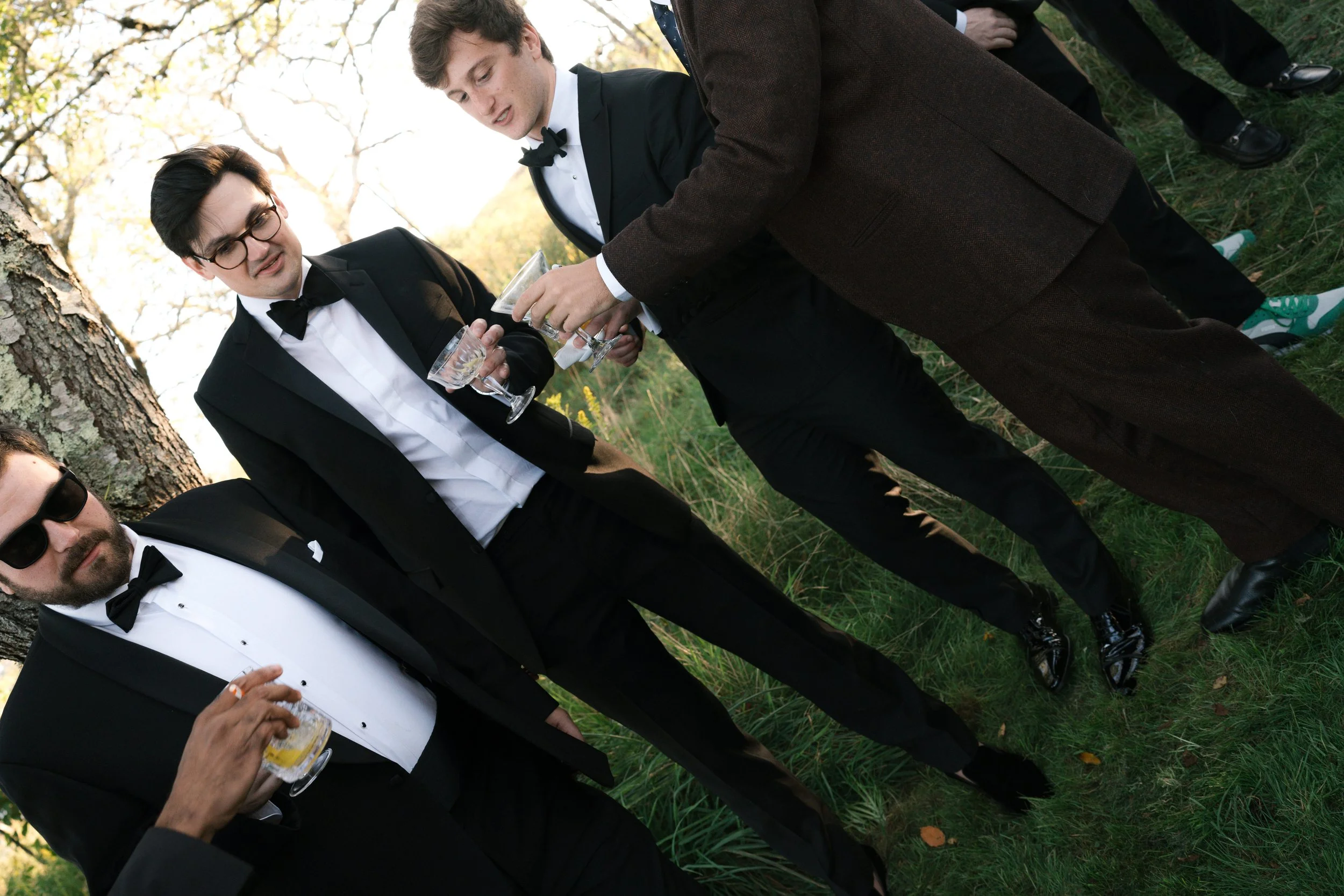 Four men in tuxedos and suits stand outdoors, holding glasses of drinks, conversing near a tree during daytime.