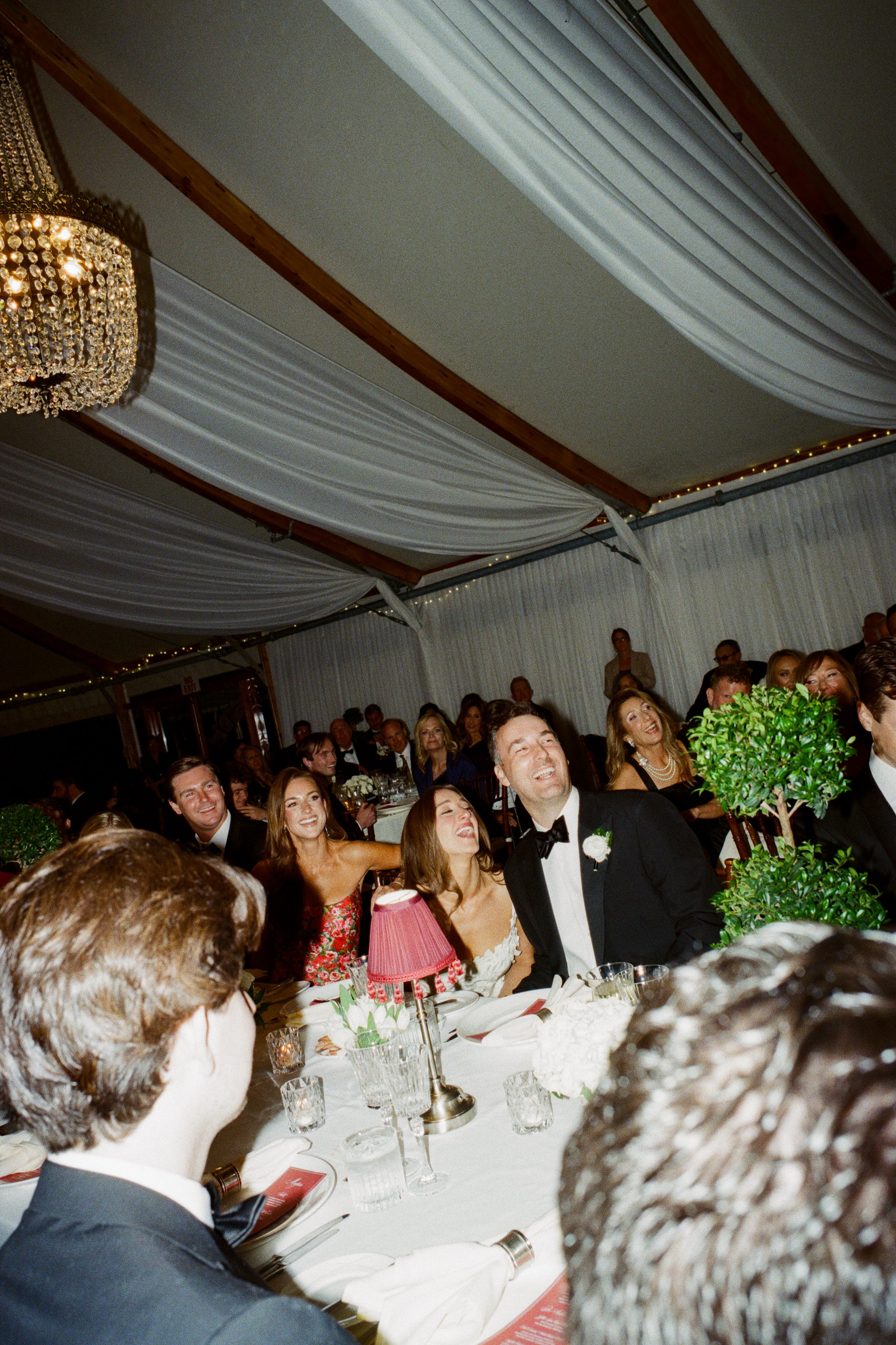 Wedding reception with guests laughing and enjoying themselves, decorated with white drapery on the ceiling and a chandelier.