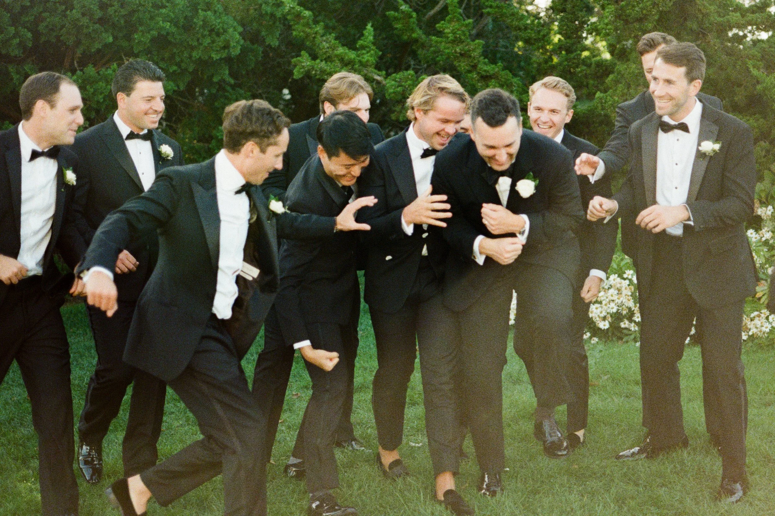 Group of men in tuxedos, laughing and playfully wrestling outdoors on green grass, surrounded by trees and bushes.