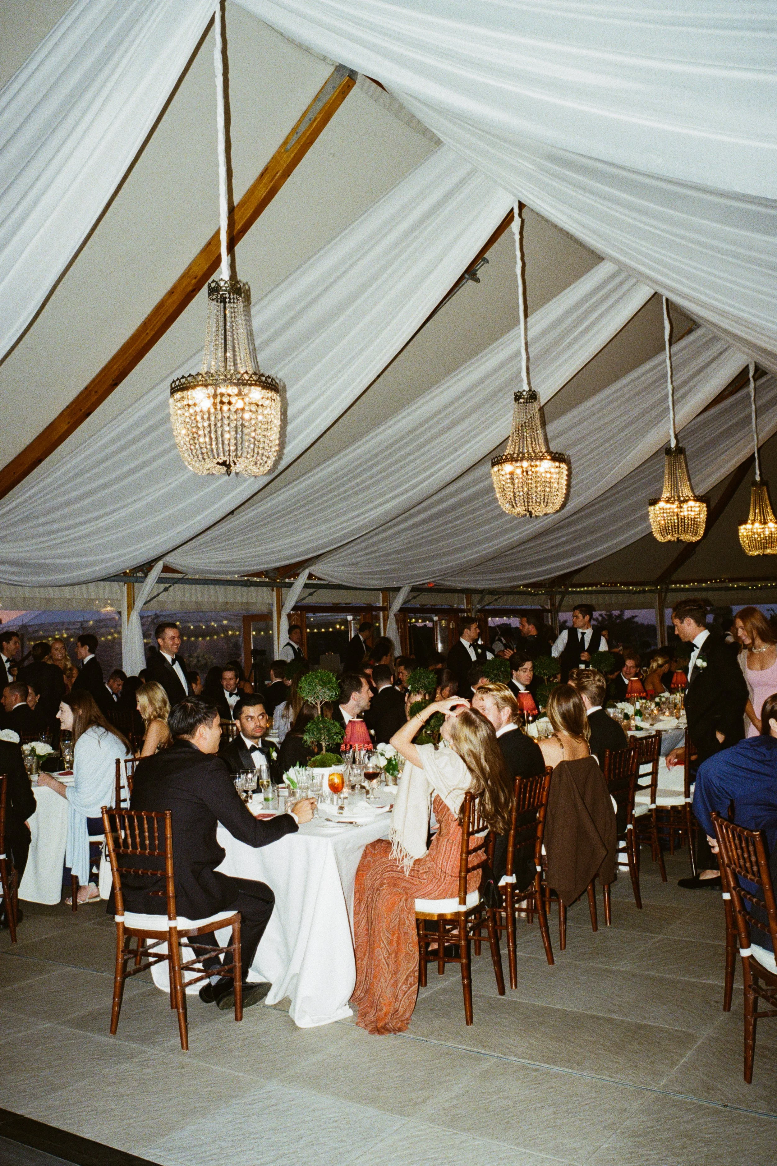 Guests at a formal wedding reception inside a decorated tent, sitting and standing around tables with white tablecloths, floral centerpieces, and red lamps, under a draped ceiling with chandeliers.