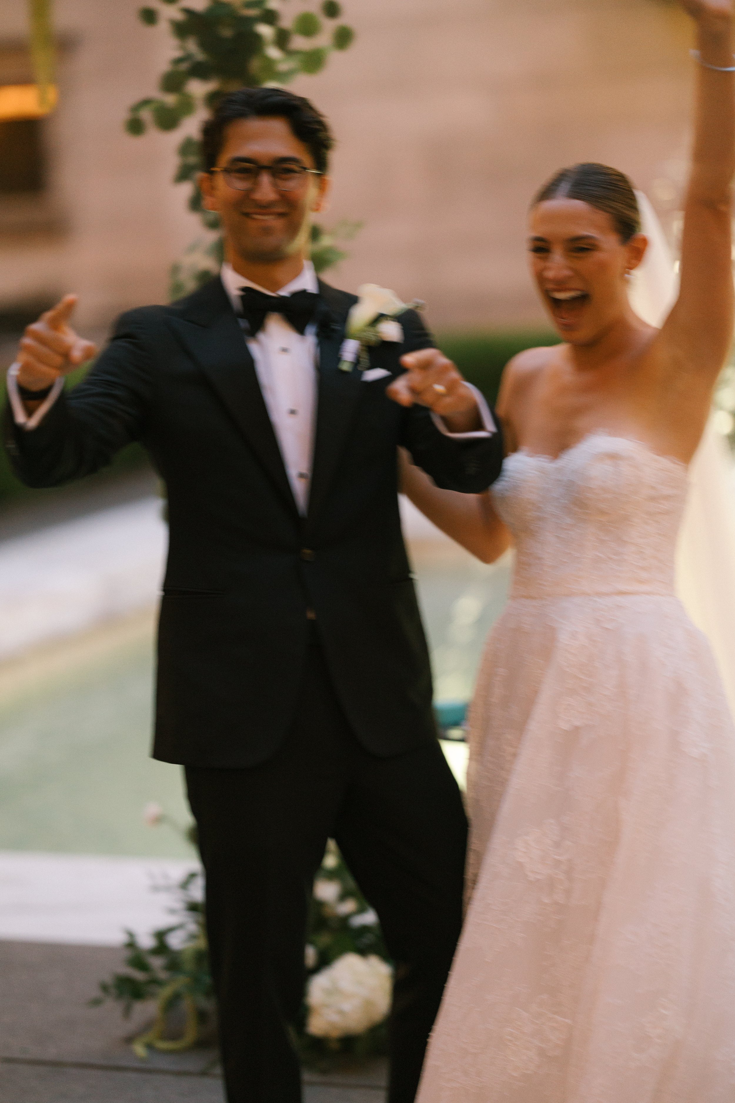 A newlywed couple in wedding attire celebrating and smiling, with the bride raising her arm and both pointing upwards.