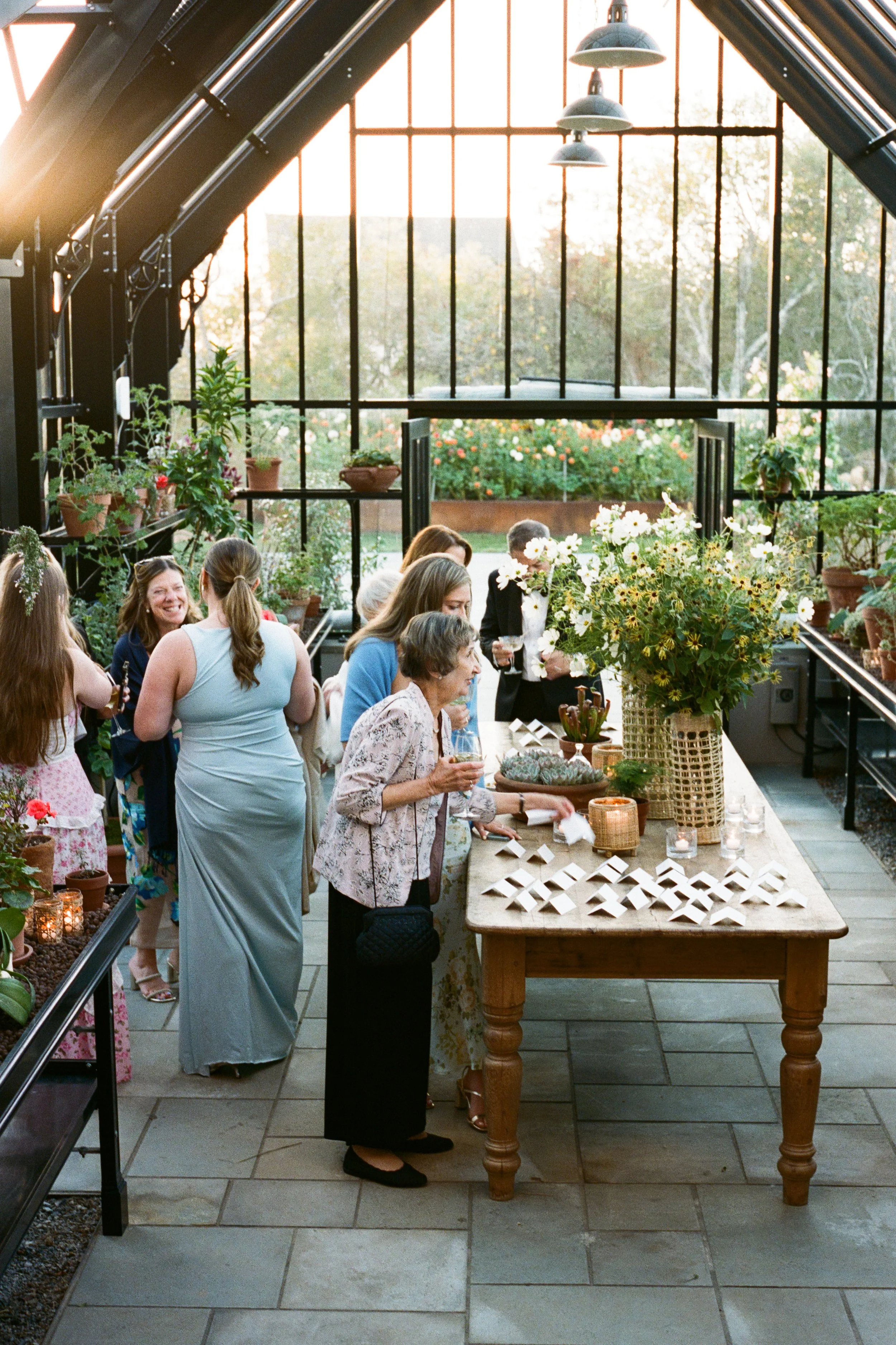 People gathered in a greenhouse with large windows, enjoying a social event around a table decorated with flowers and candles.