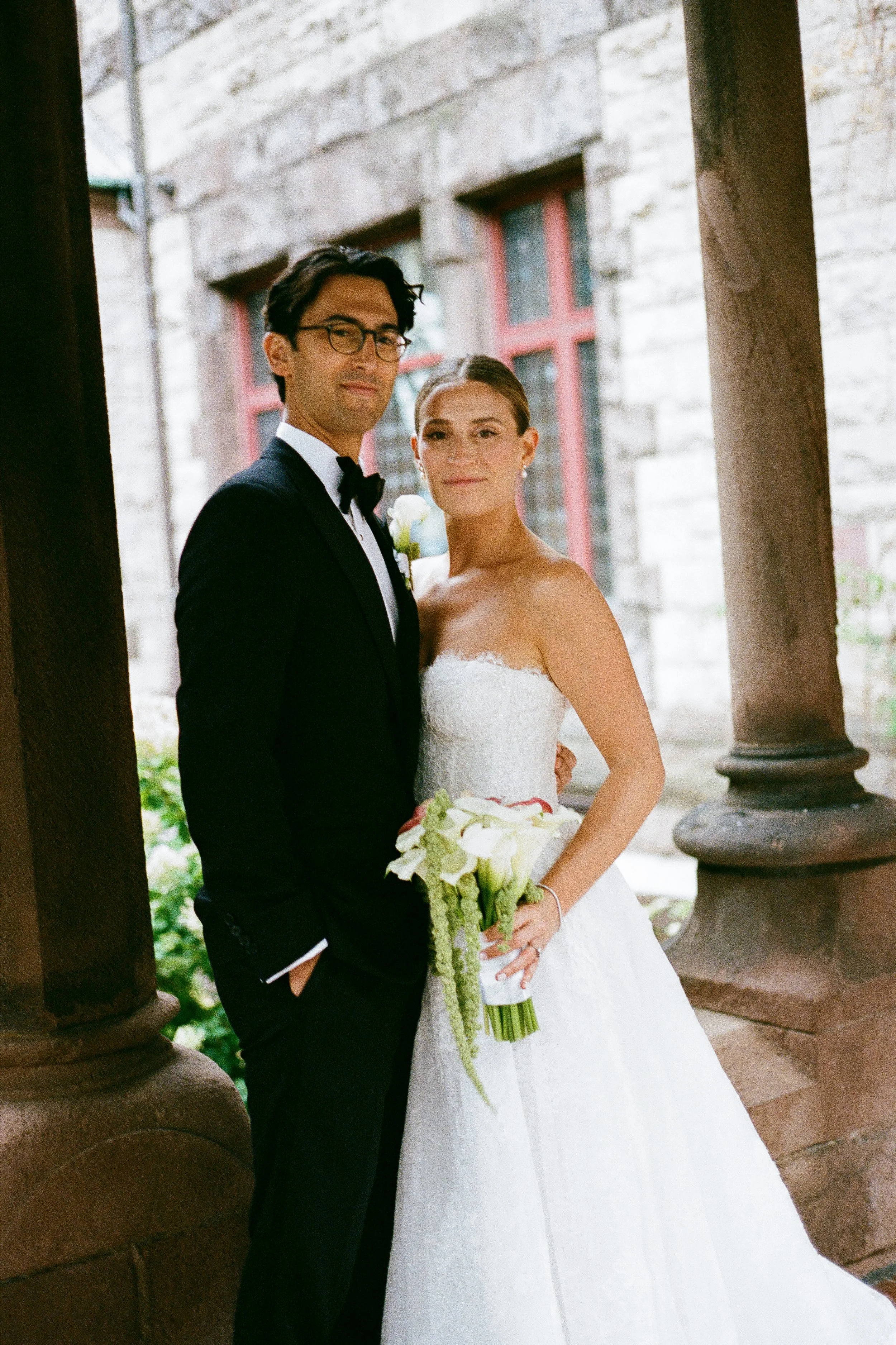 Bride and groom in wedding attire standing under stone columns.