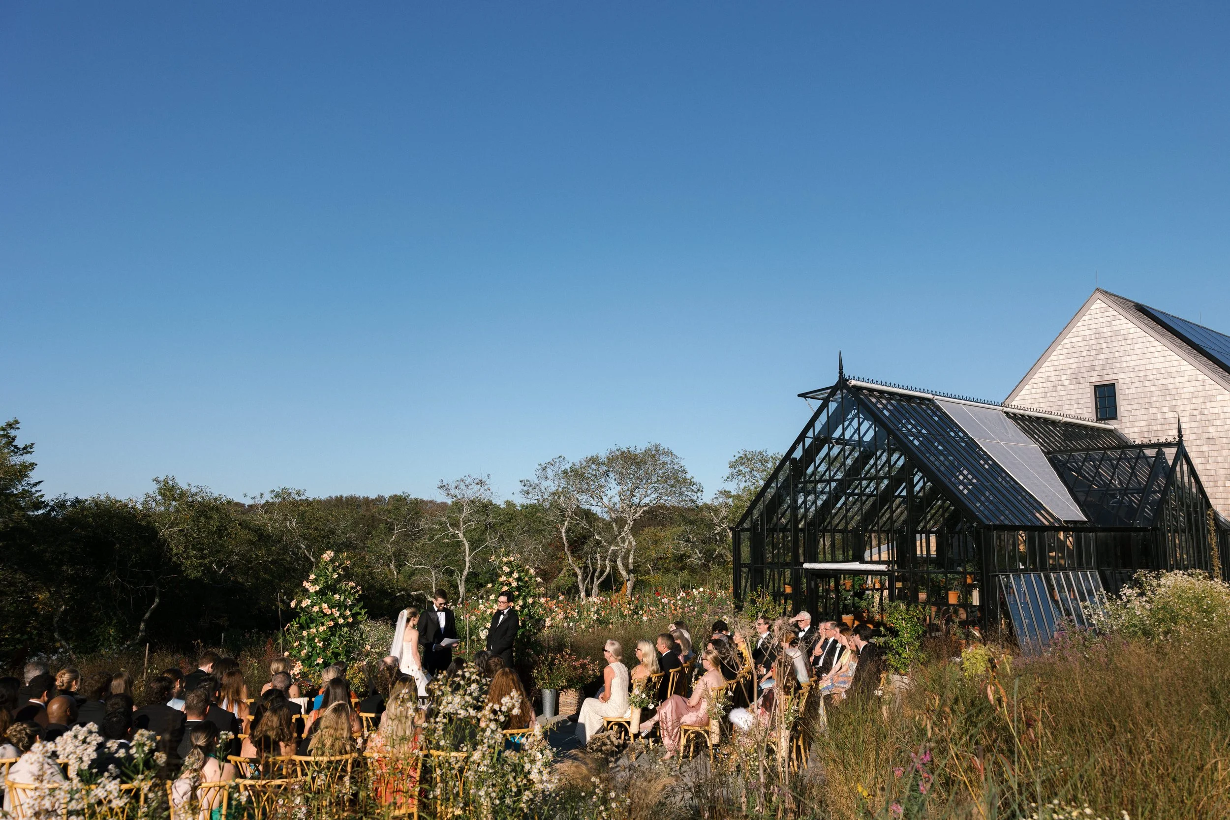 Outdoor wedding ceremony with guests seated in front of a glass greenhouse and a white barn under a clear blue sky.