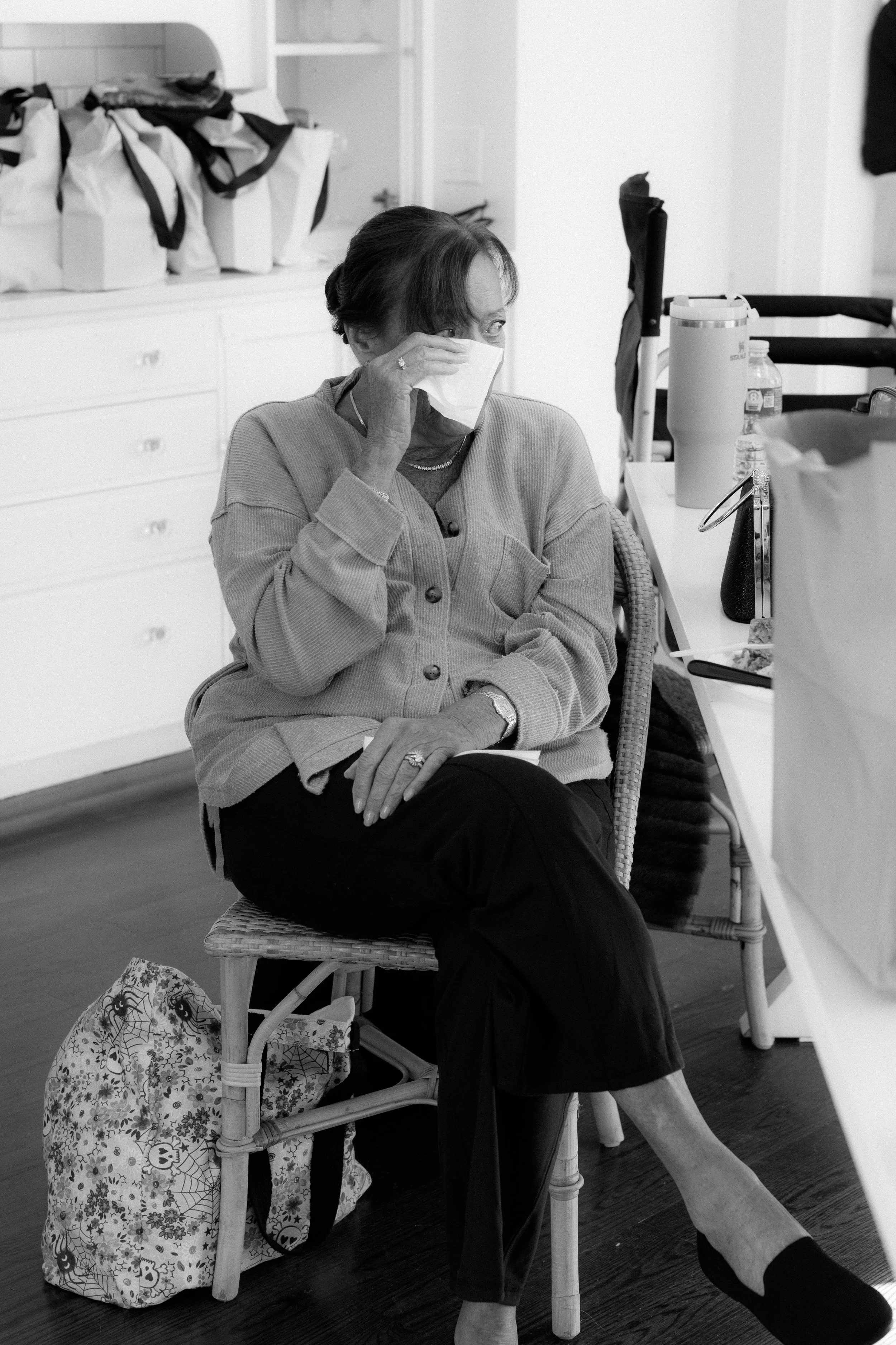 An elderly woman sitting on a wicker chair, wearing a face mask, grey cardigan, black pants, and black shoes, is in a room with a white cabinet and various bags on top. She seems to be adjusting her mask or preparing to speak.