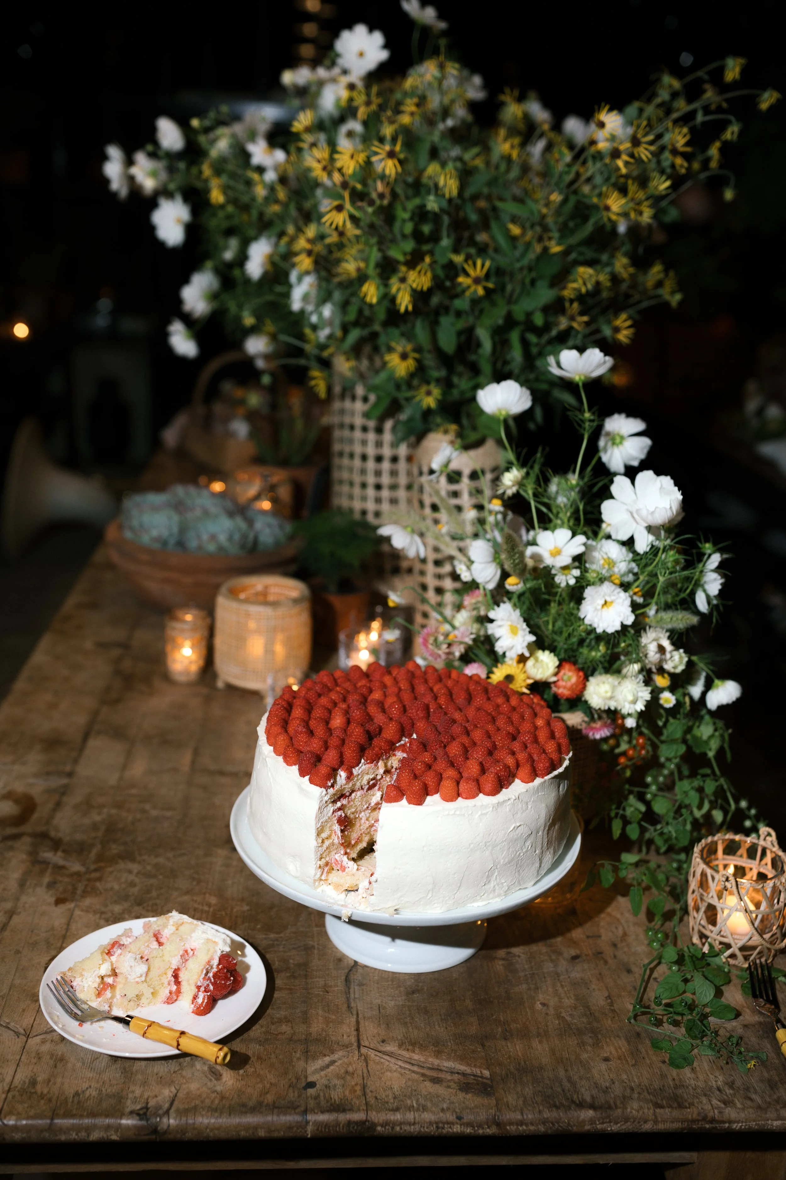 A partially sliced white cake topped with raspberries on a cake stand. There is a plate with a fork and a piece of cake in the foreground. The background has a rustic wooden table with flowers and candles.
