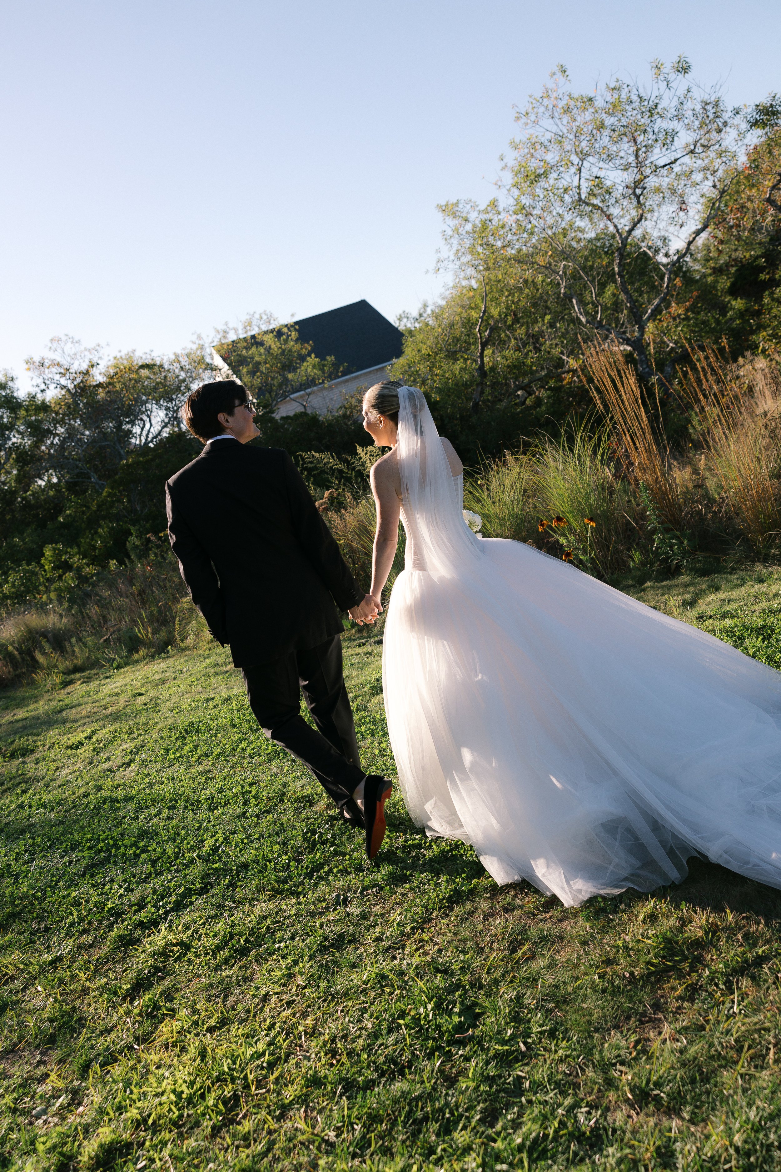A bride and groom holding hands and walking outside on a grassy area, with trees and a house in the background, during golden hour.