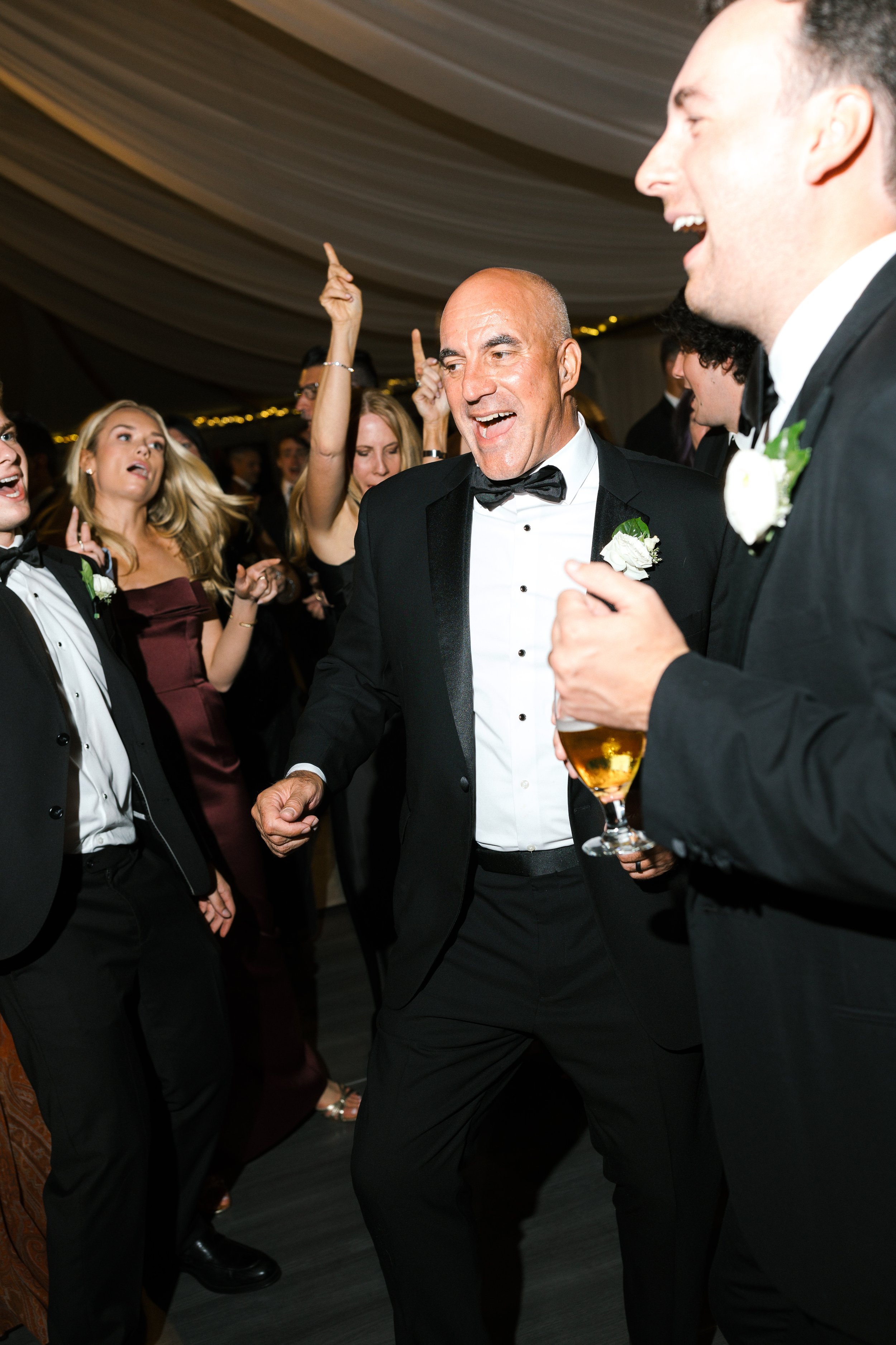 Men and women dancing and celebrating at a formal event or wedding reception, with party attire and drinks in hand.