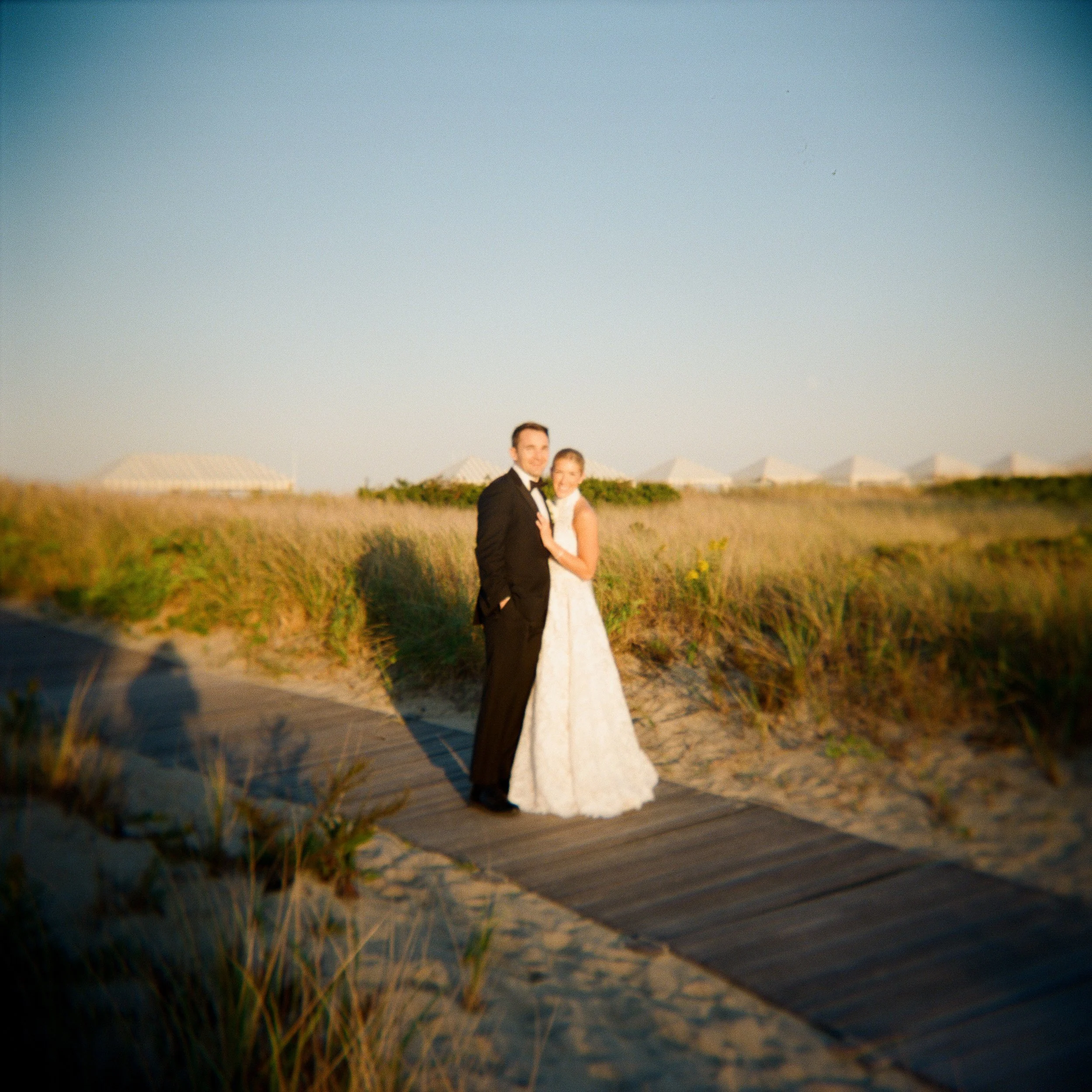 A bride and groom in wedding attire standing on a sandy path with tall grass and seaside structures in the background during sunset.
