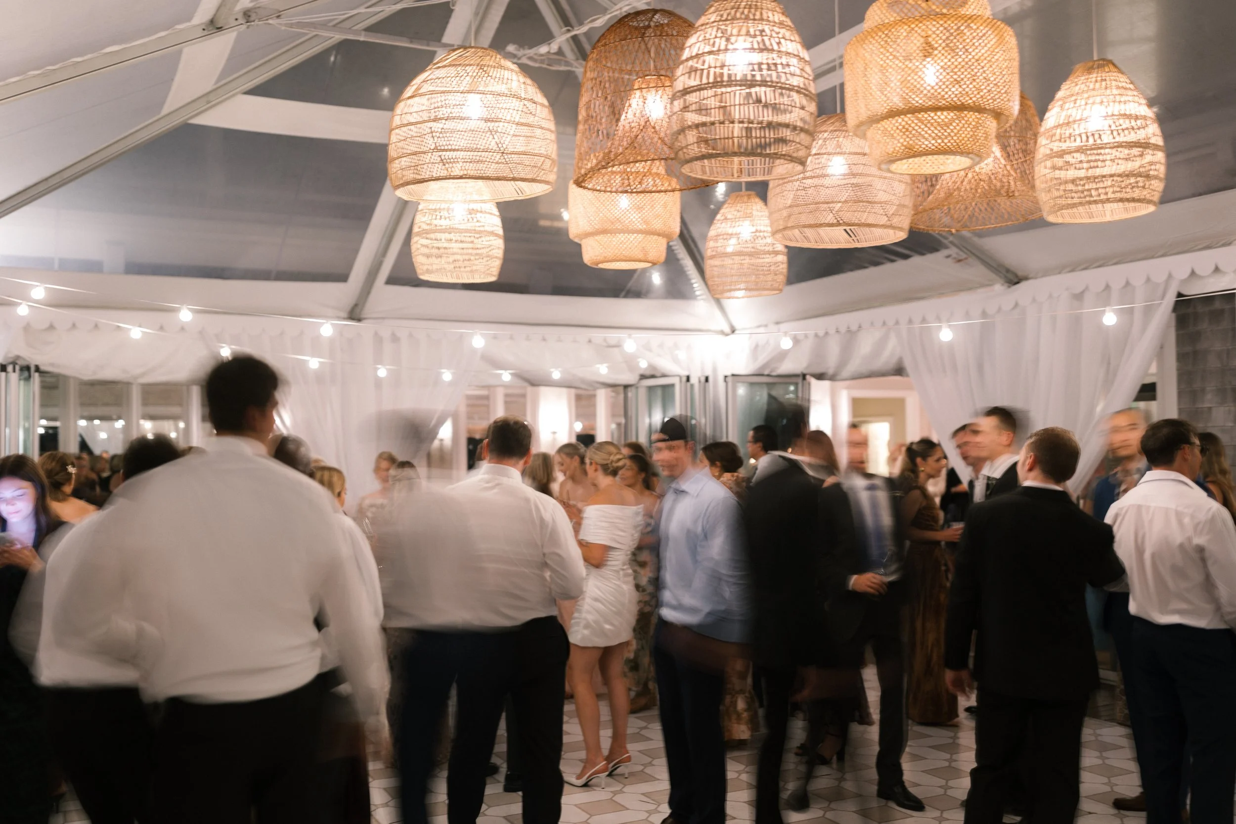 People dancing and socializing at an indoor wedding reception with hanging wicker lights and white drapery.