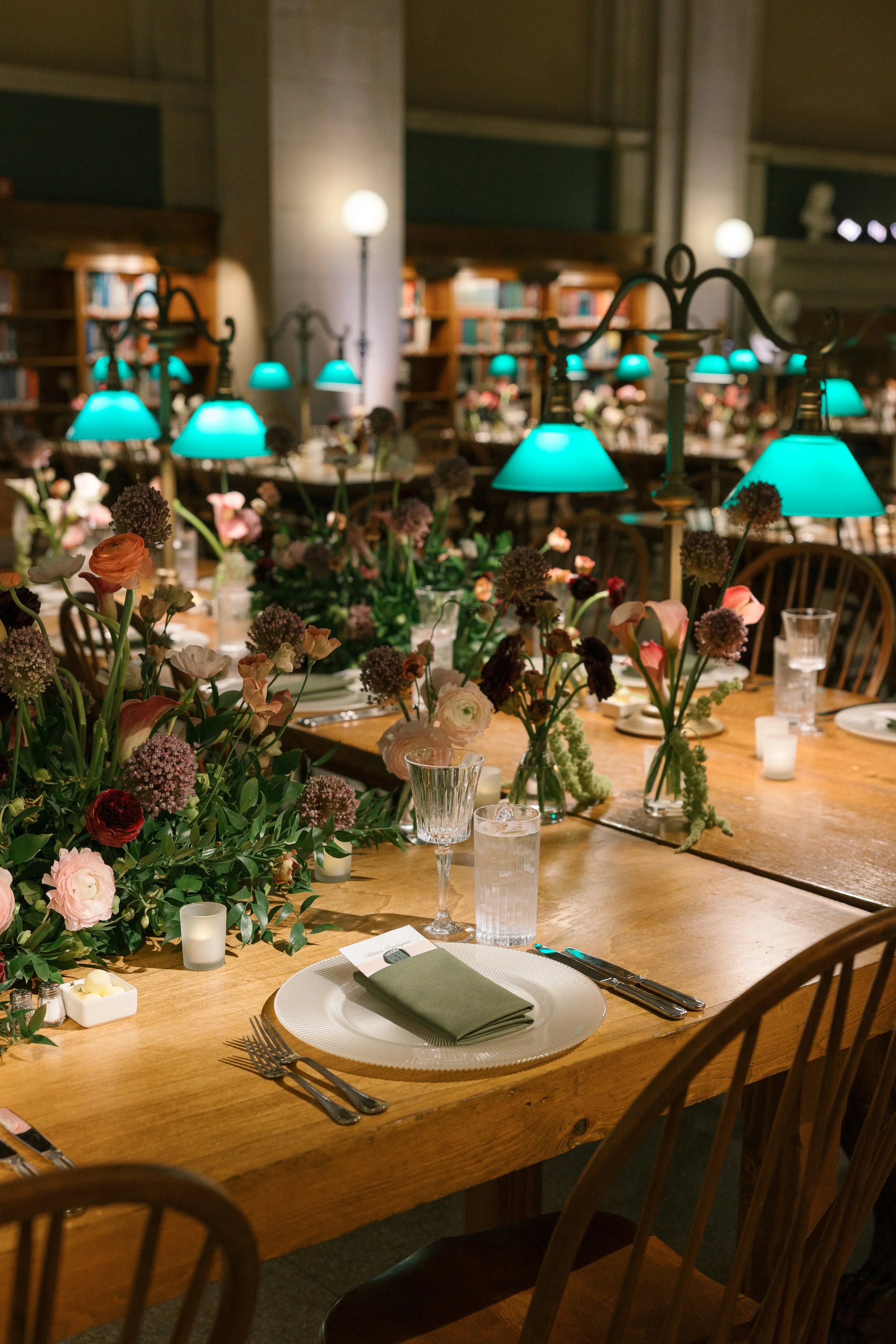 A decorated dining table with floral centerpieces, place settings, and teal pendant lights in a library or restaurant setting.