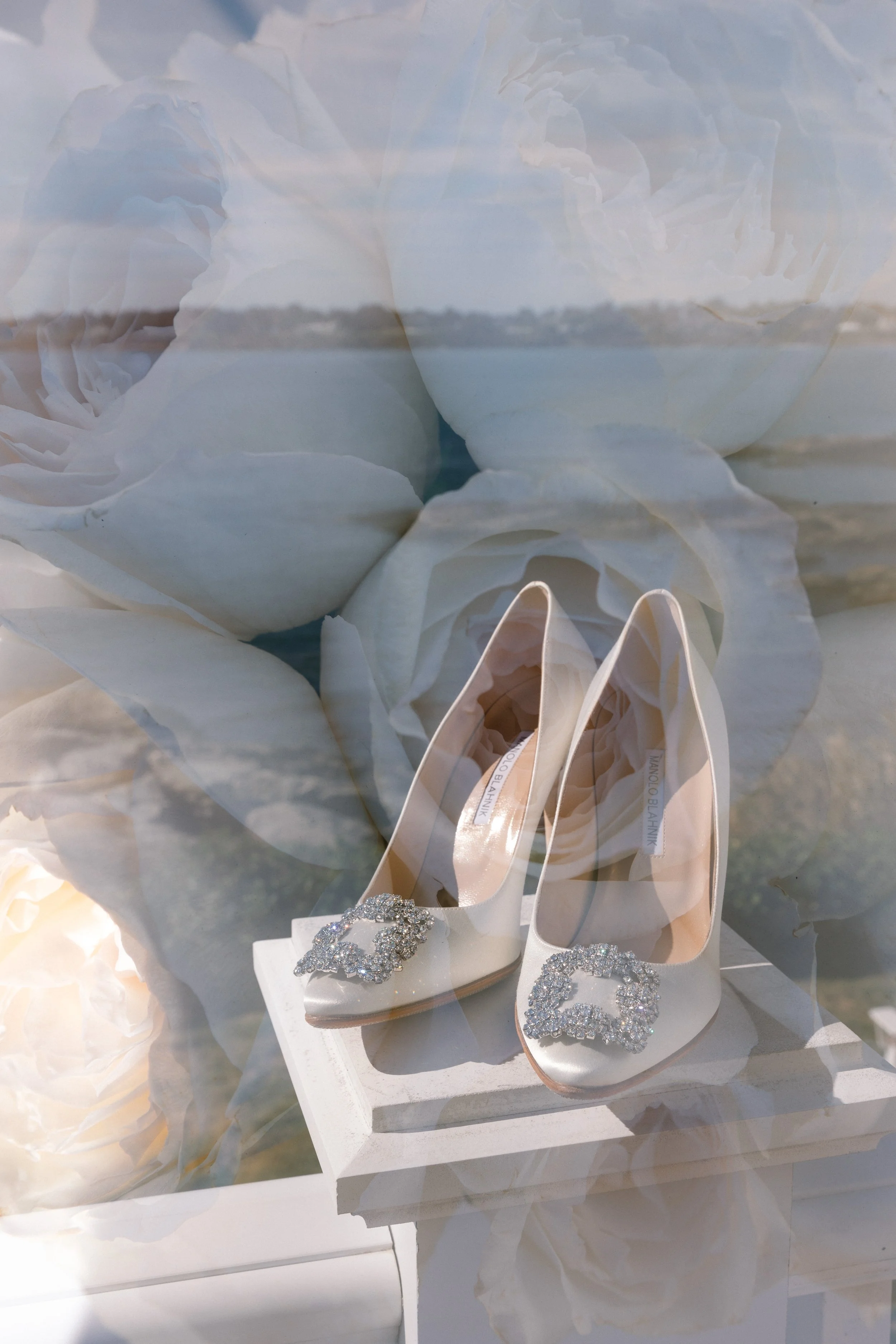 A pair of white high heels with large rhinestone embellishments on the toe area, placed on a white surface with a background featuring large white roses.