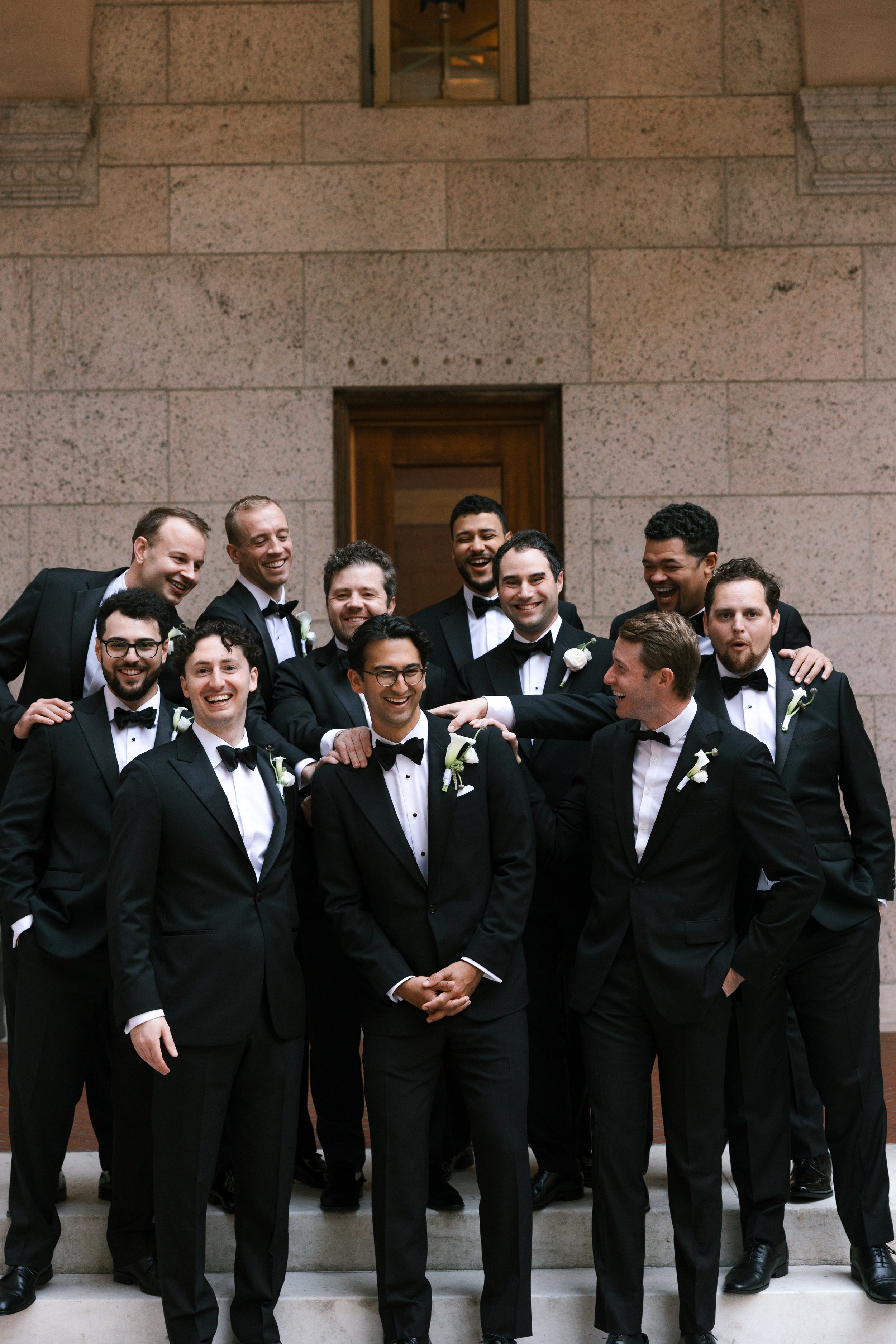 A group of men dressed in black tuxedos with bow ties, standing on steps in front of a stone building, smiling and posing for a photo at a wedding.