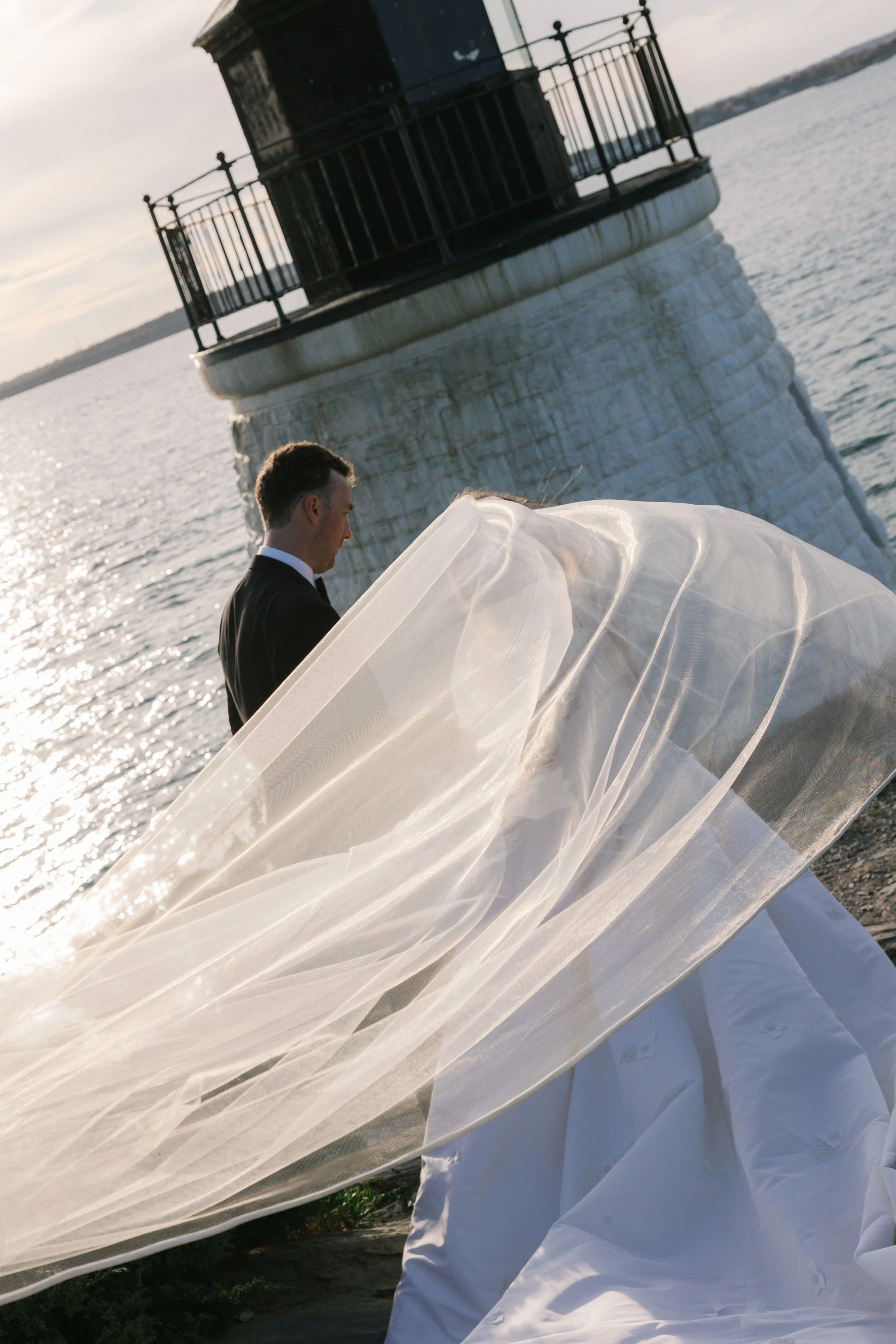 A man in a dark suit stands outdoors near water, with a white wedding gown and veil draped over a surface in the foreground. There is a lighthouse in the background.