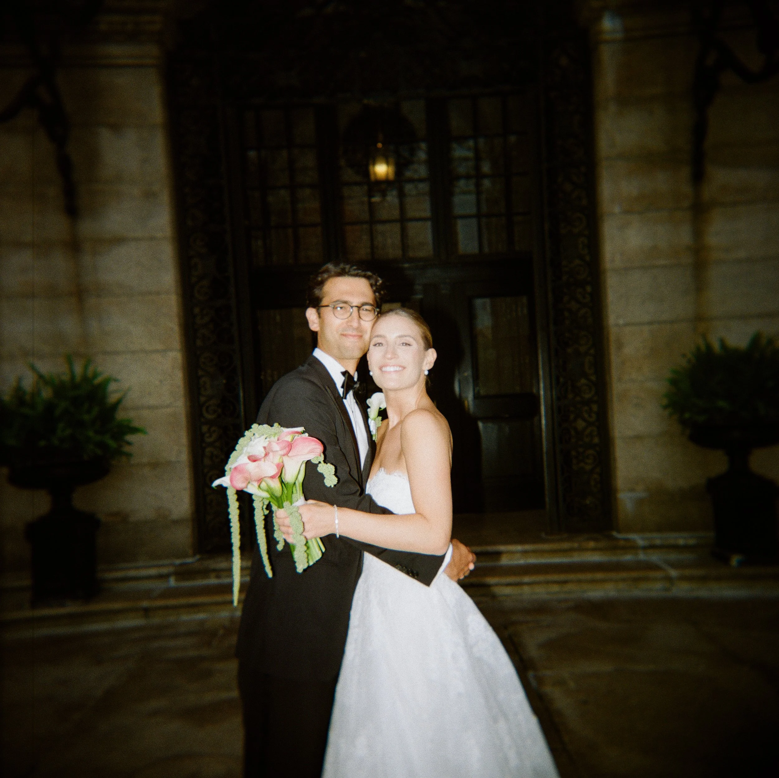 A bride and groom standing together outside a building, smiling at the camera. The groom is wearing a black tuxedo with a bow tie, and the bride is in a white strapless wedding gown holding a bouquet of pink and white flowers. The background features